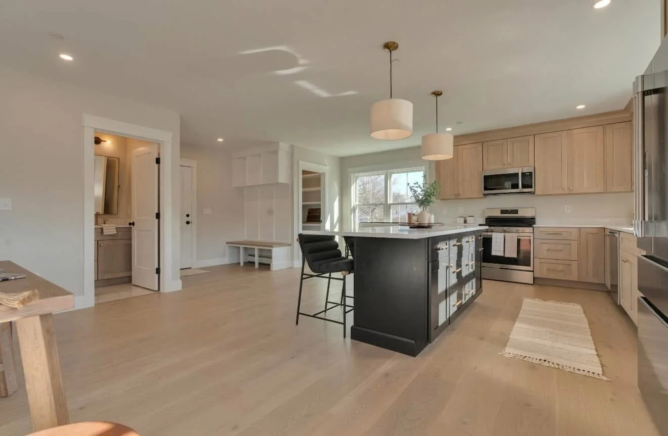 Modern kitchen with light wood cabinets, stainless steel appliances, black kitchen island, ceiling lights, and a view of the living area and dining space.