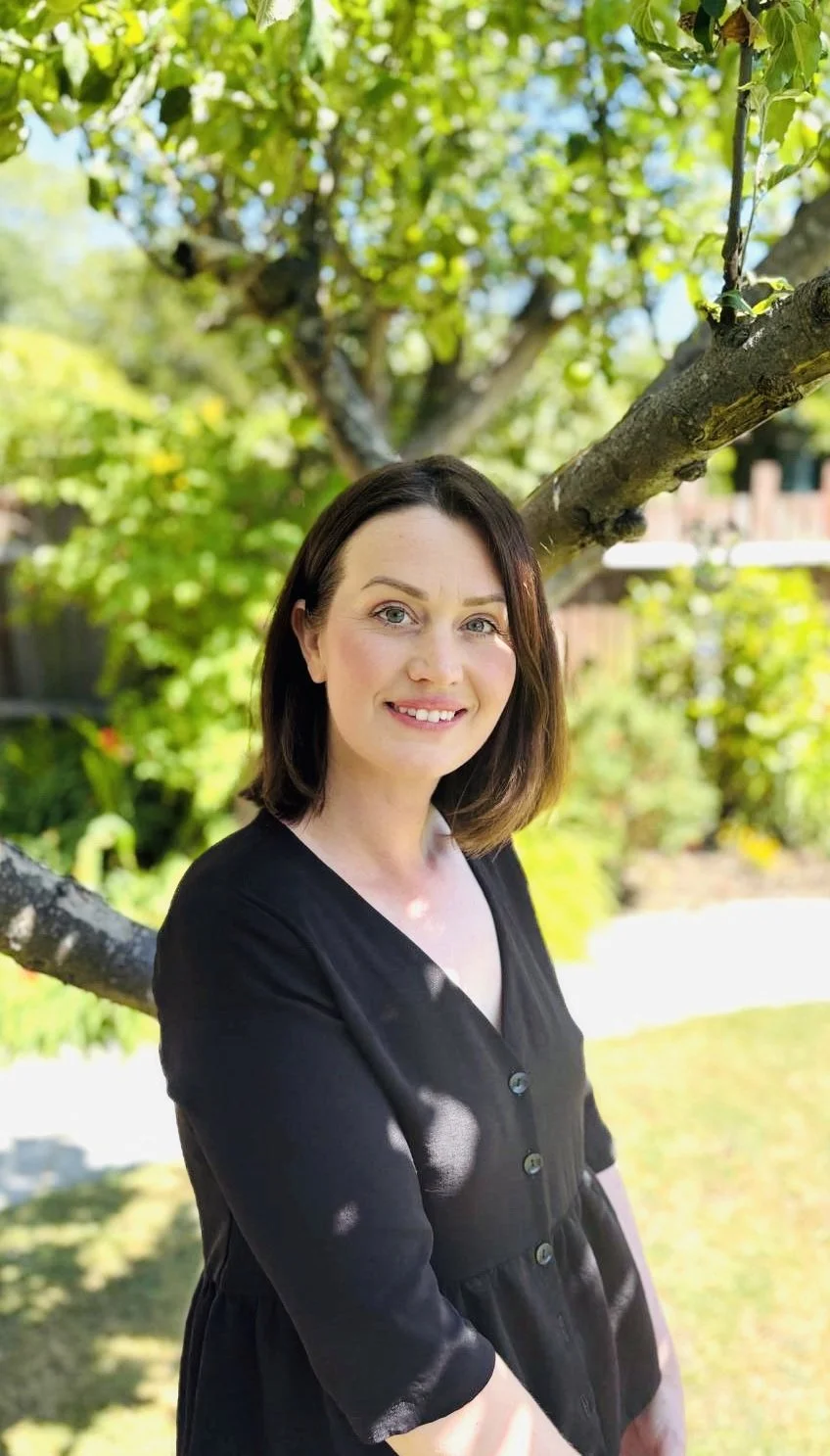 A woman with shoulder-length dark hair and blue eyes standing outdoors in a garden under a tree with green leaves.