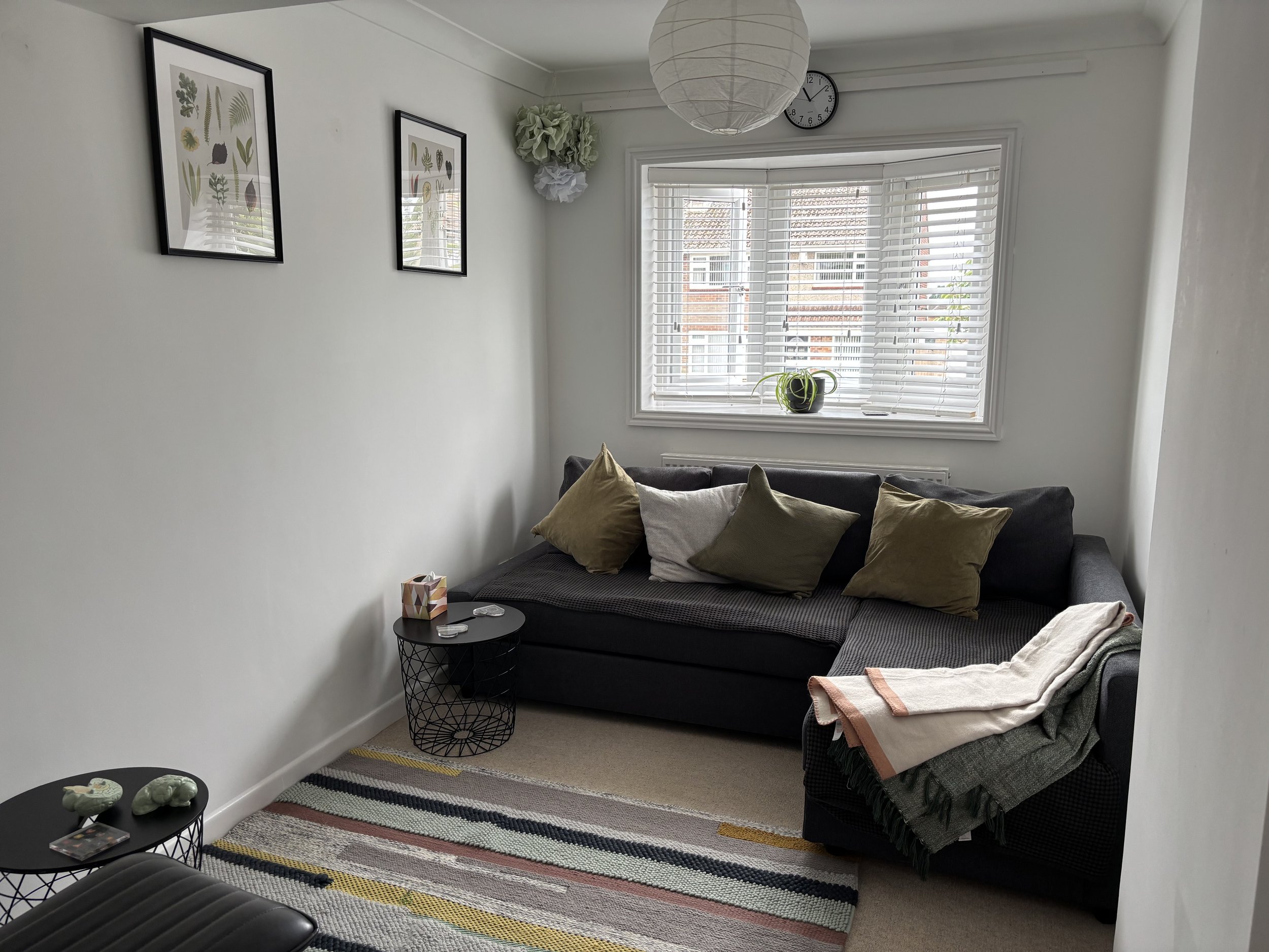 A small living room with white walls and a large window with white blinds. There is a dark gray sofa with four pillows and a blanket. Two round black wire side tables with items on top are in front of the sofa. A striped rug is on the beige carpeted floor. Two framed botanical art prints hang on the wall, and a wall clock and paper lantern light fixture are visible.