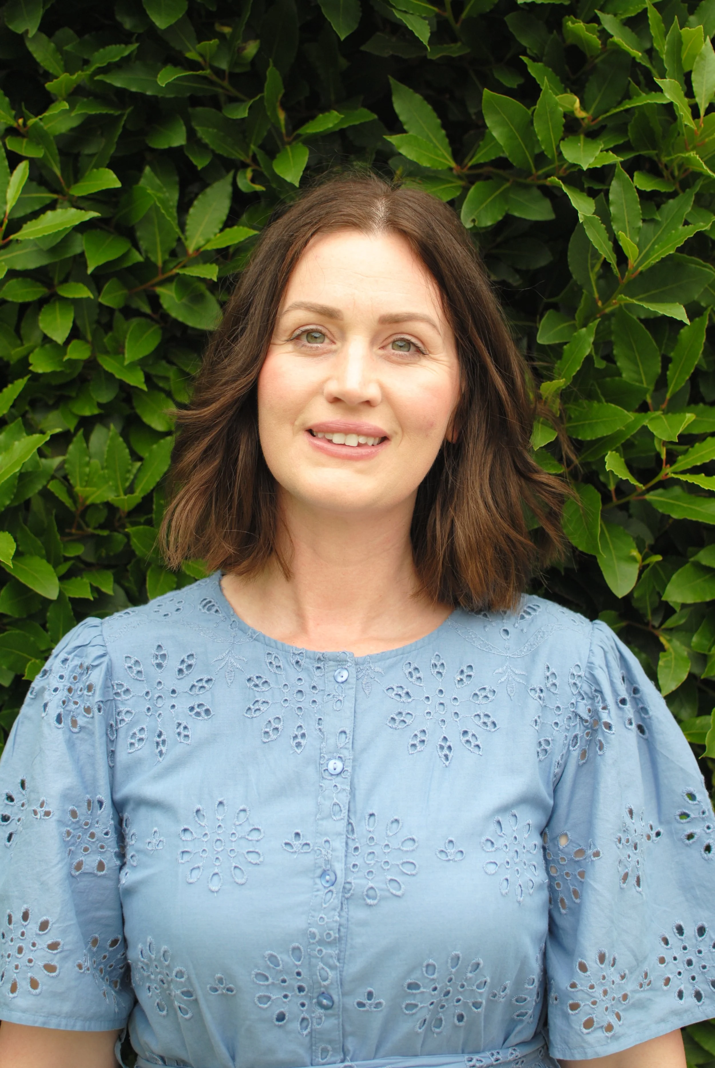 A woman with shoulder-length brown hair and green eyes smiling in front of green foliage, wearing a light blue blouse with floral eyelet embroidery.
