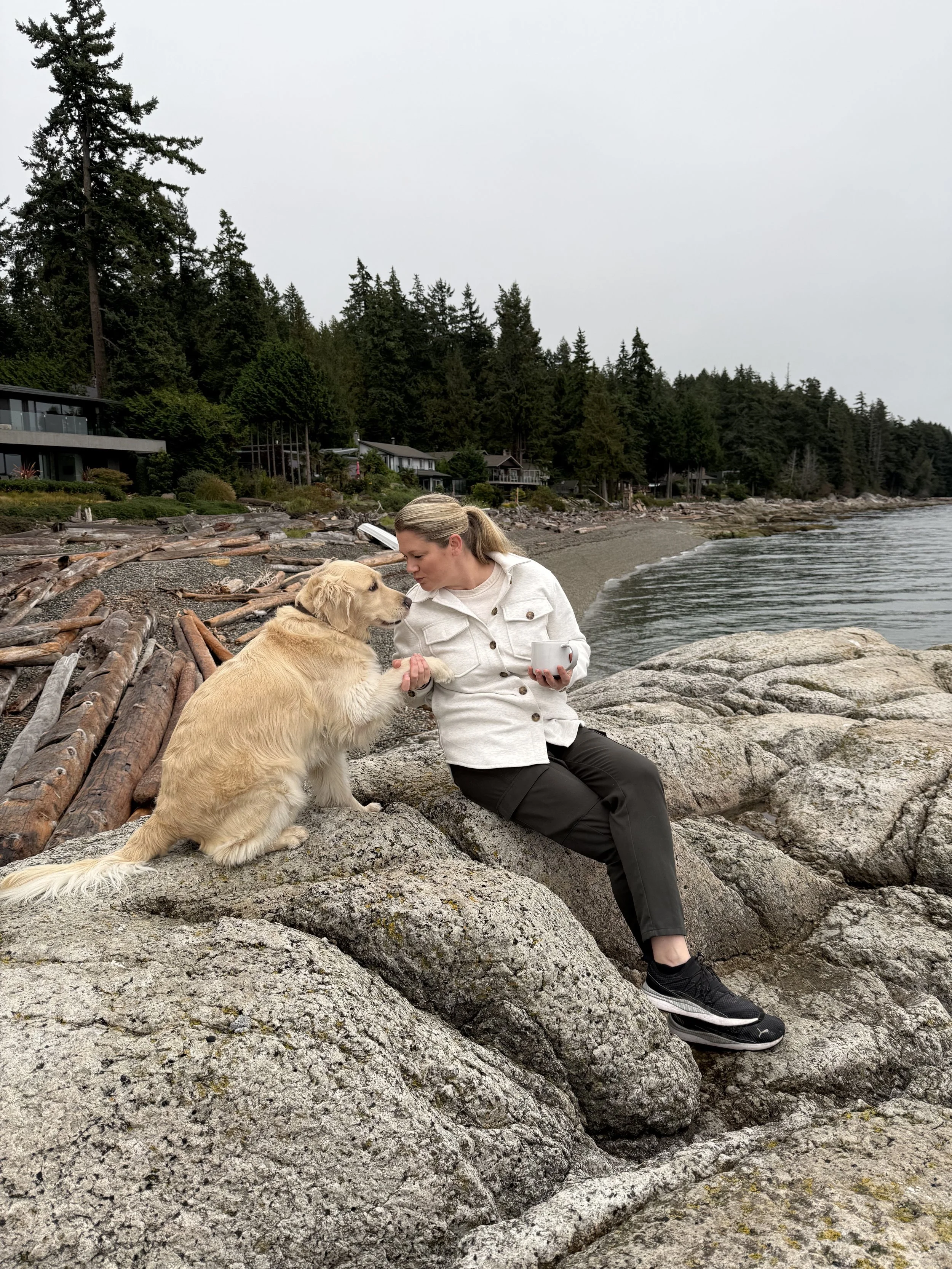 A woman sitting on rocks by the water, holding a smartphone, with a dog sitting beside her on a rocky shoreline amidst trees and residential houses.