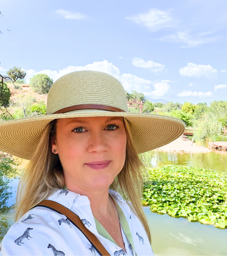 A woman wearing a wide-brimmed straw hat and a white shirt with zebra print, standing outdoors near a pond with lily pads, trees, and a blue sky with clouds in the background.