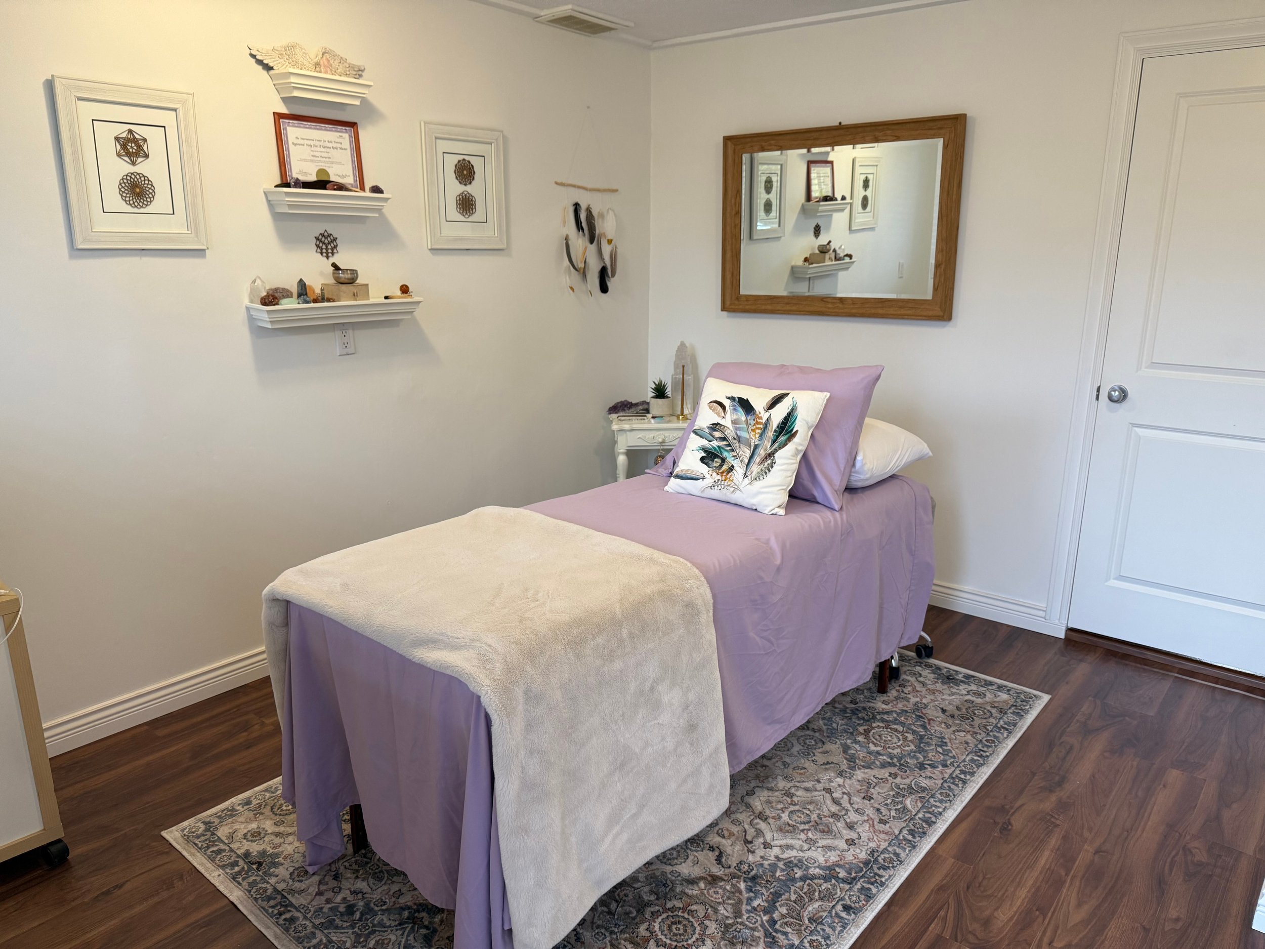A small room with a massage or therapy bed covered with a purple sheet and a white blanket. There is a decorative pillow with feathers on it and a white pillow behind it. The room has a patterned area rug on wooden floor, a white wall with framed artwork and shelves, a mirror on the wall, and a door.