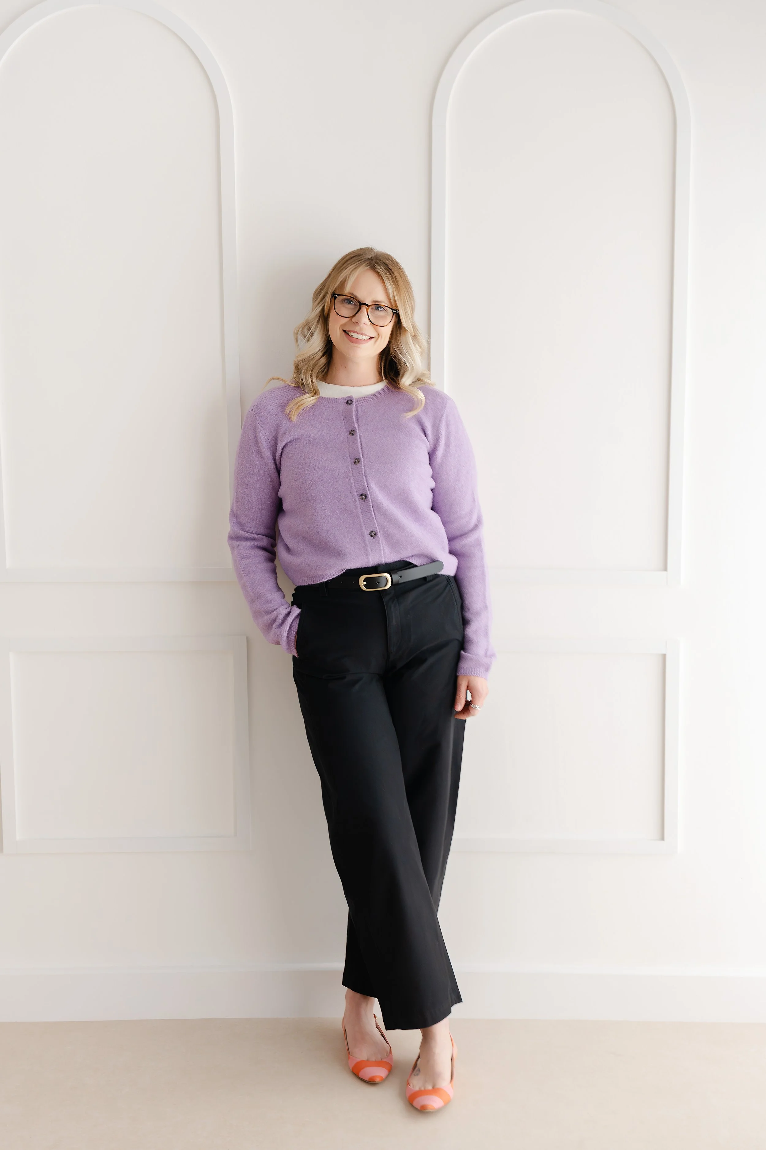 A woman standing in front of a white wall, wearing a lilac cardigan, black trousers with a belt, and peach and pink flat shoes. She has blonde, wavy hair and is wearing glasses, smiling at the camera. The woman is a counsellor and psychotherapist.