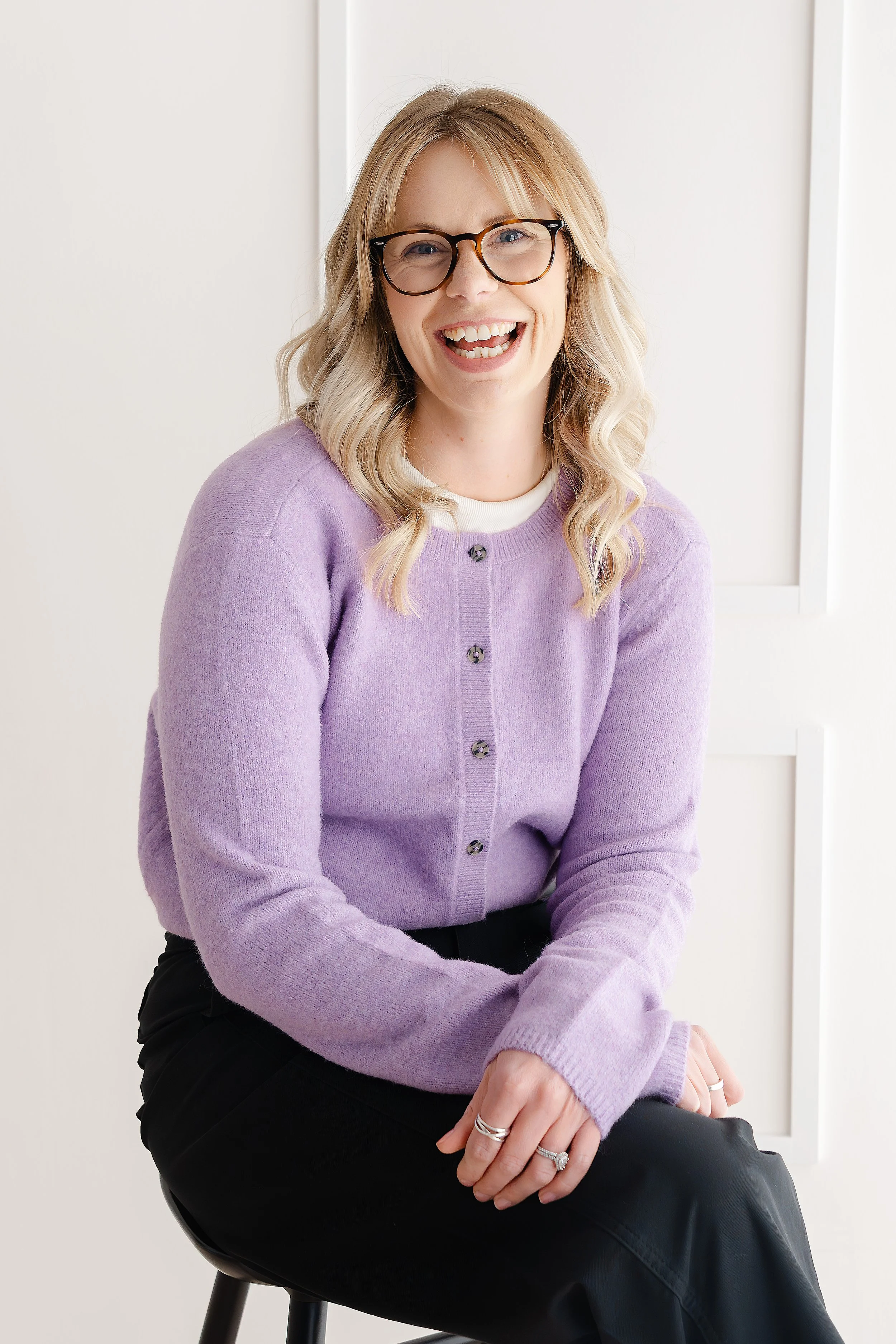 A woman with blonde hair and glasses sitting on a chair, wearing a purple cardigan and black trousers, smiling and looking at the camera. The woman is a counsellor and psychotherapist based in Yeovil, UK.