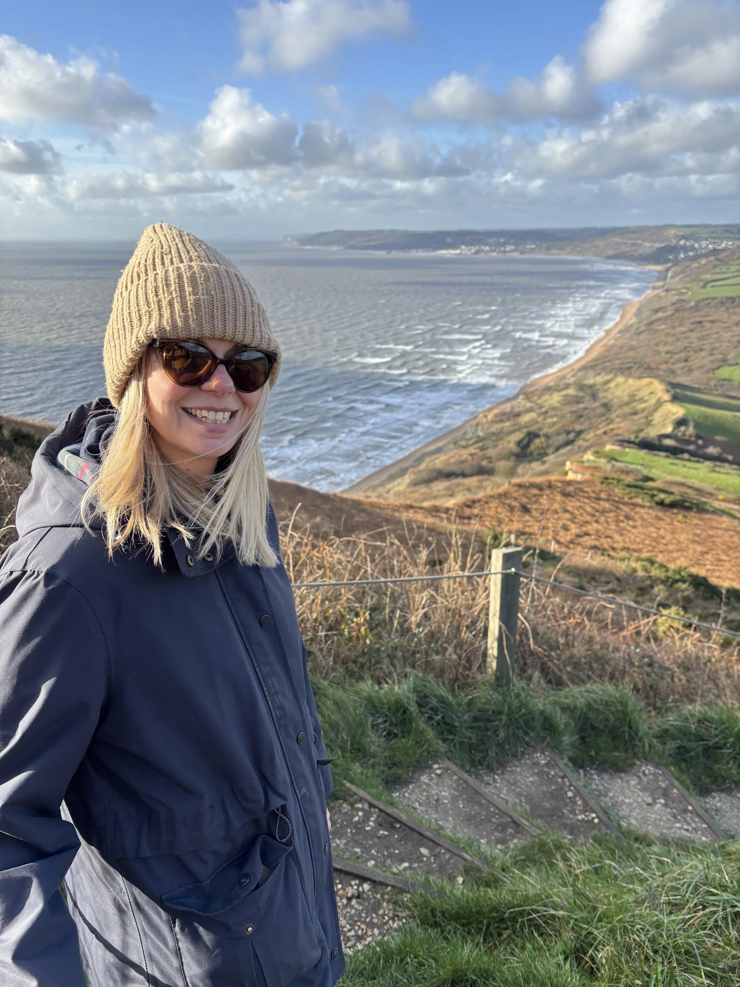 Woman in a navy jacket, tan knit hat, and sunglasses smiling outdoors with a coastline, ocean, cliffs, and hills in the background.