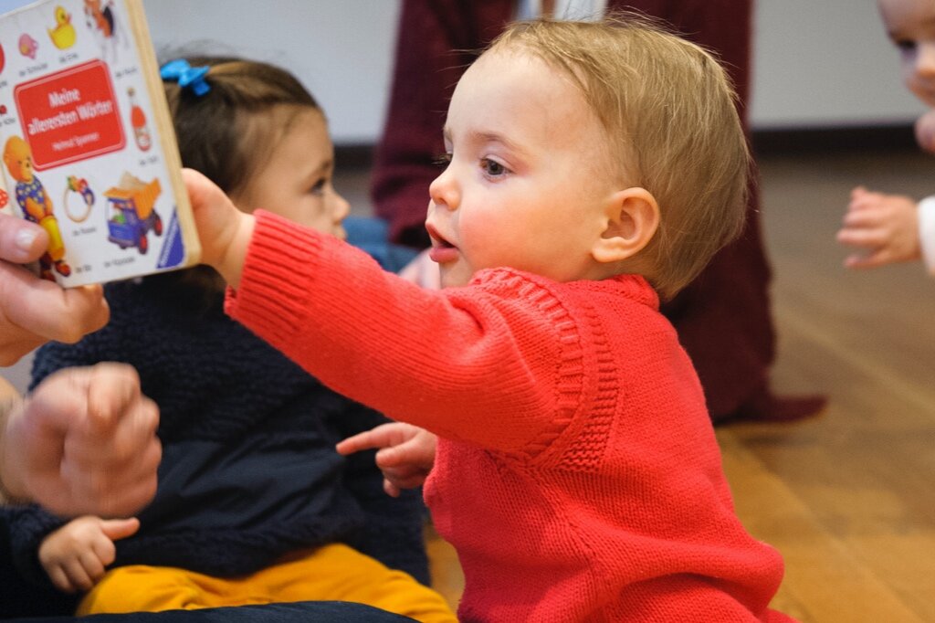 A young boy (about 18 to 24 months) reaches up to touch a book page while it is being read to him
