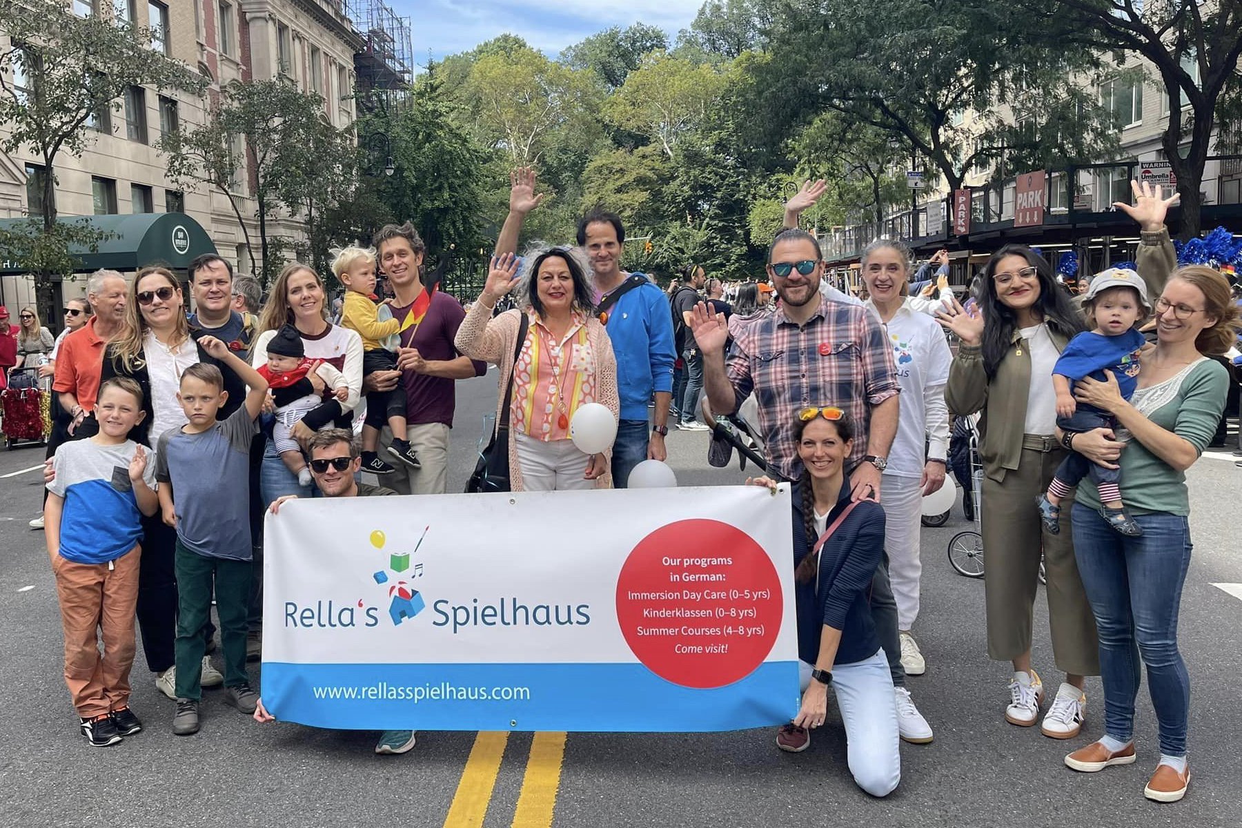 Group of Rella's Spielhaus parents and their children, teachers,  and friends waving "Guten Tage" while two people are holding a banner.