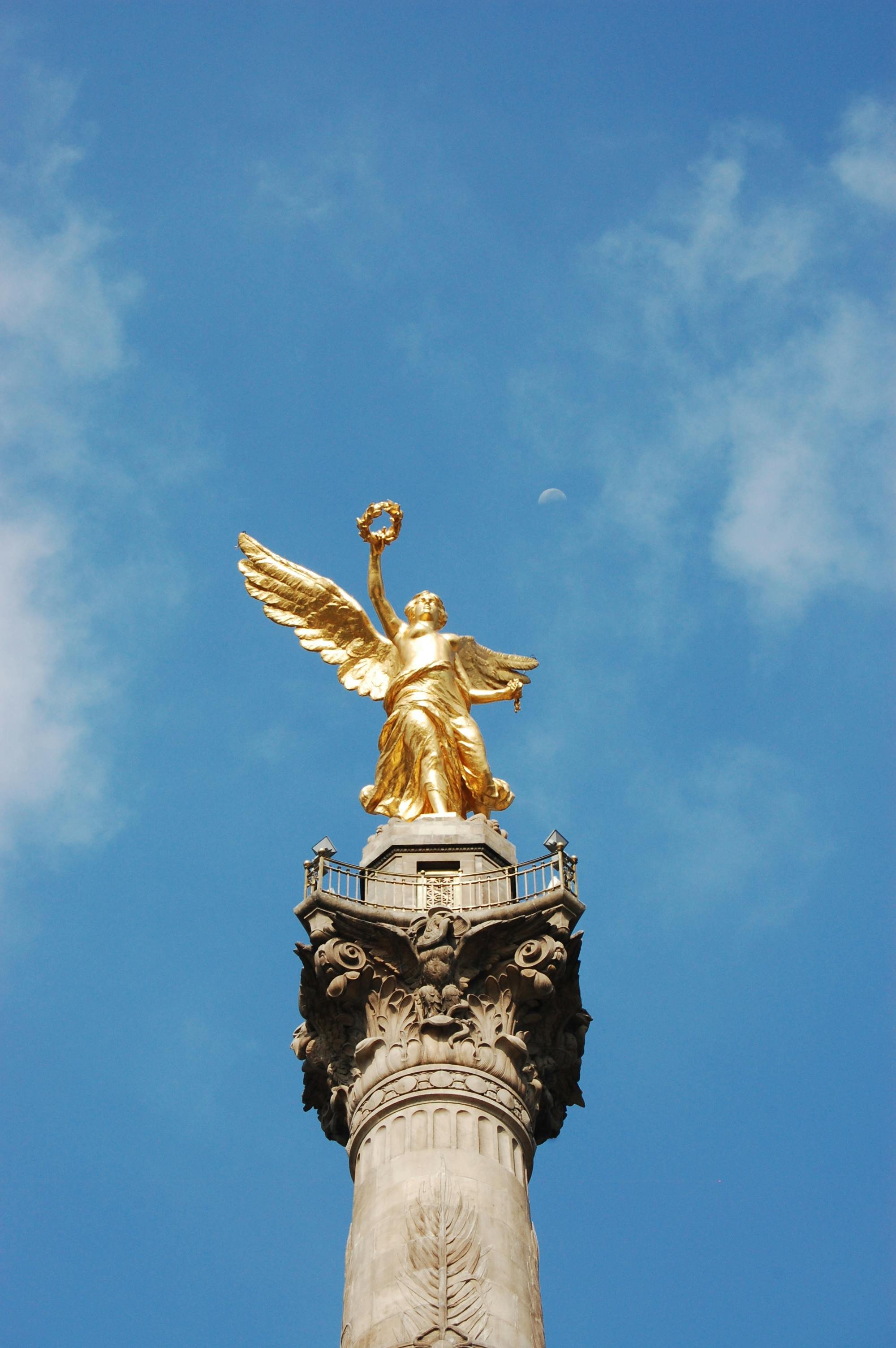 Golden statue of a winged female figure holding a wreath, atop an ornate column against a blue sky with clouds and a visible moon.