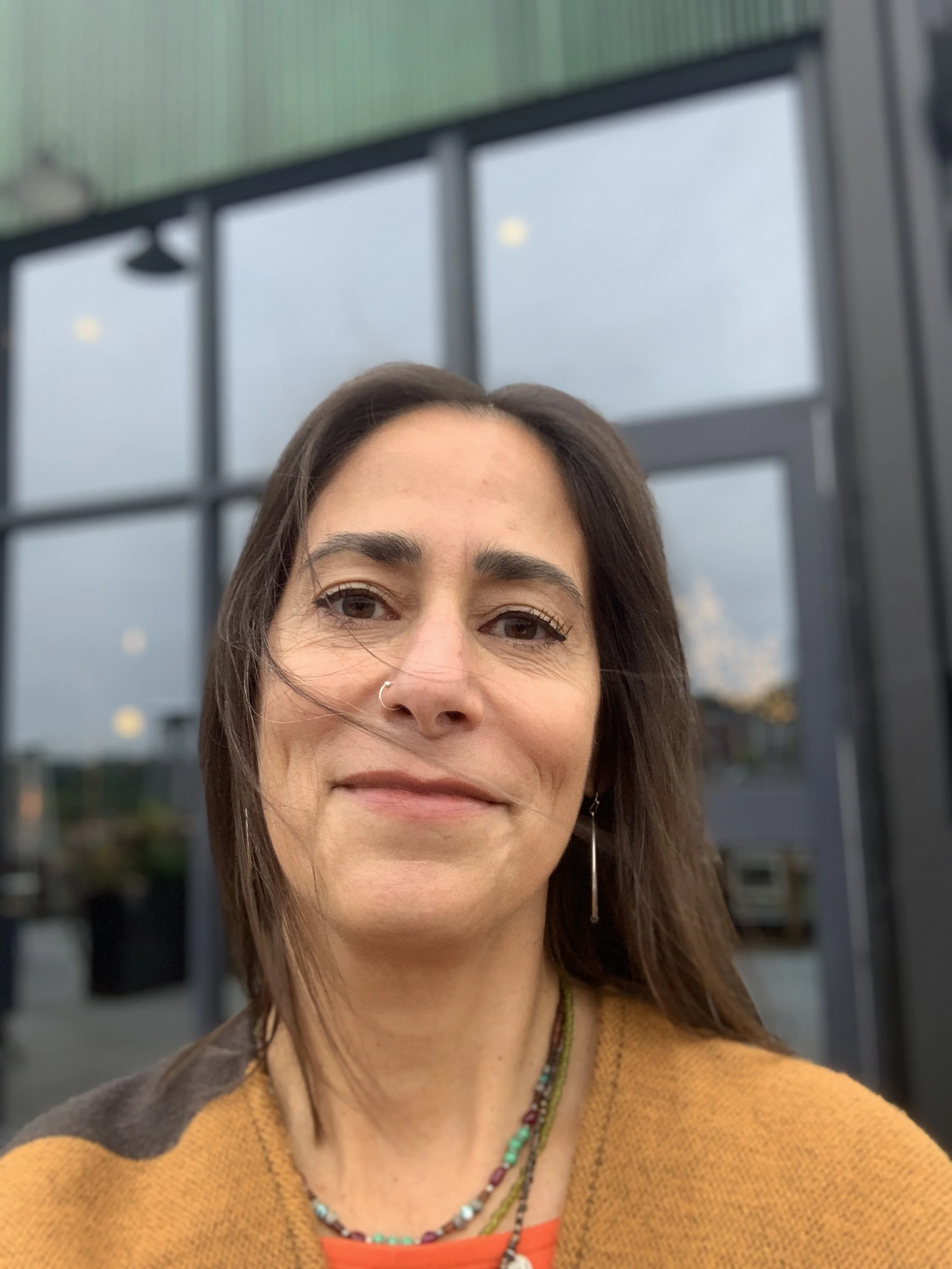 A woman with dark hair and a nose ring smiling outdoors in front of a large windowed building.