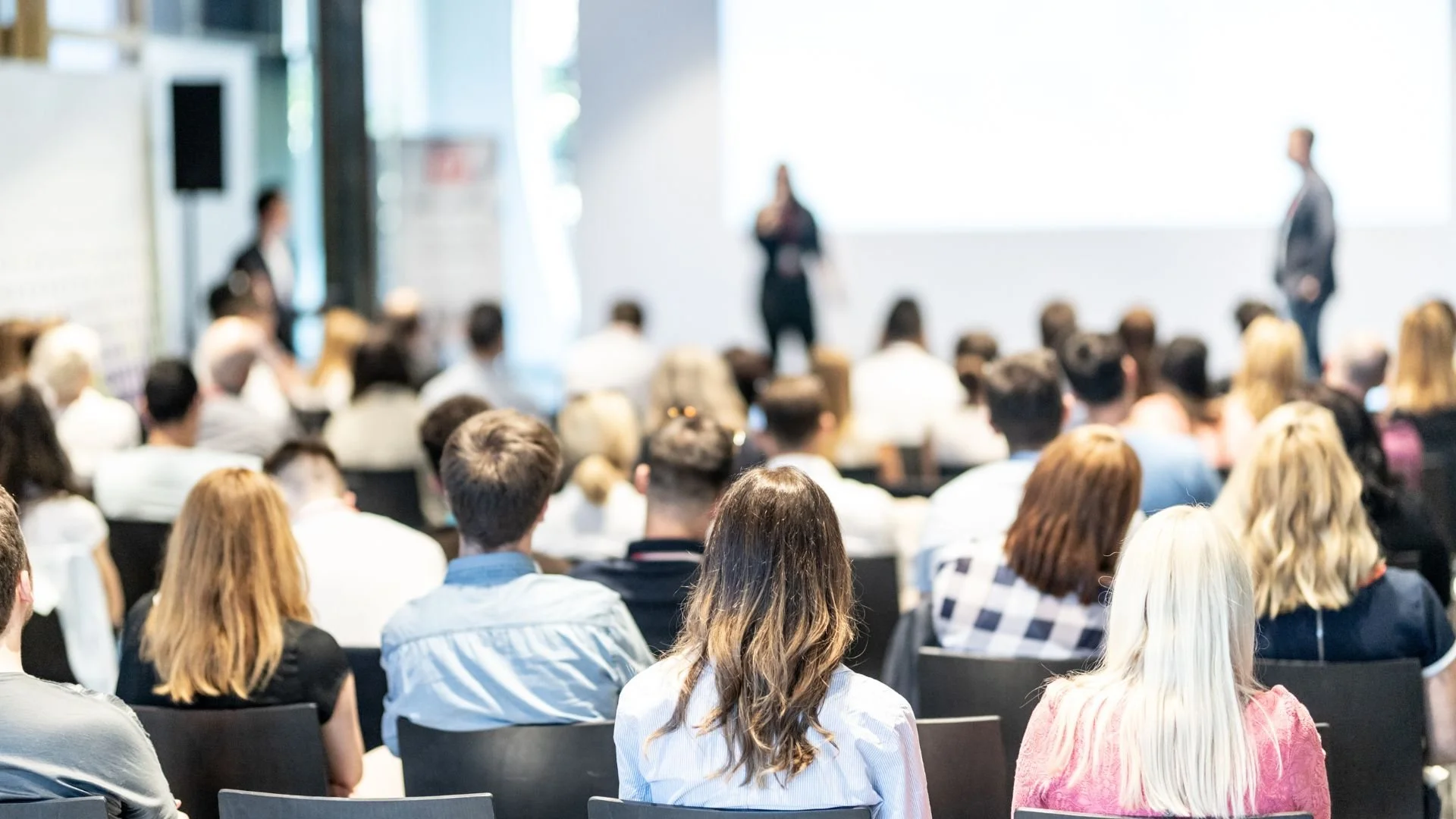 Menschen bei einer Konferenz oder Schulung im Saal, mit zwei Sprechern auf der Bühne, im Hintergrund eine große weiße Tafel oder Leinwand.