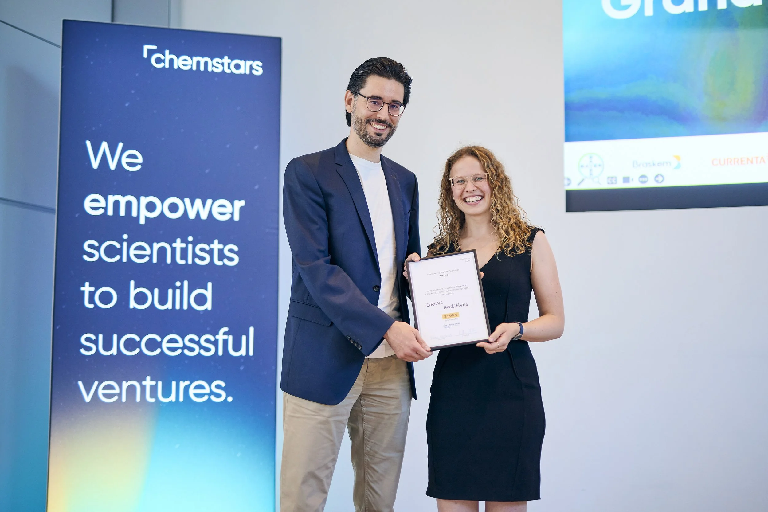 Zwei Menschen bei einer Preisverleihung, einer hält ein Zertifikat, im Hintergrund ein Bildschirm und ein Banner mit dem Text 'Chemstars' und einer Slogan."