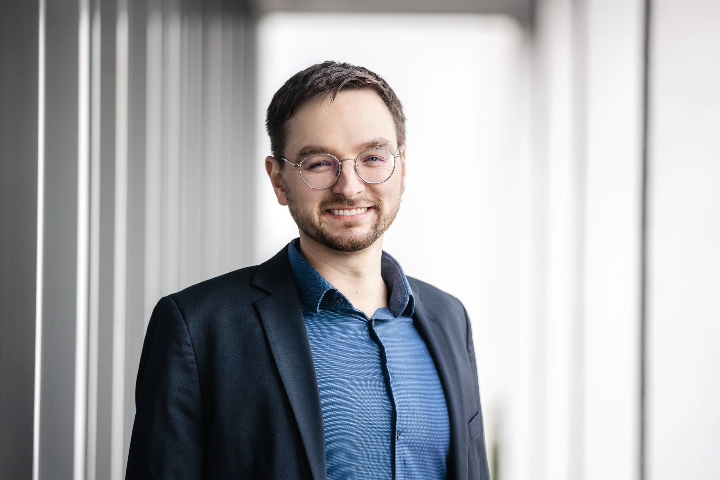 Ein junger Mann mit Brille trägt einen dunkelblauen Anzug und ein hellblaues Hemd, steht vor einer modernen Wand mit vertikalen Linien, lächelt und blickt direkt in die Kamera.
