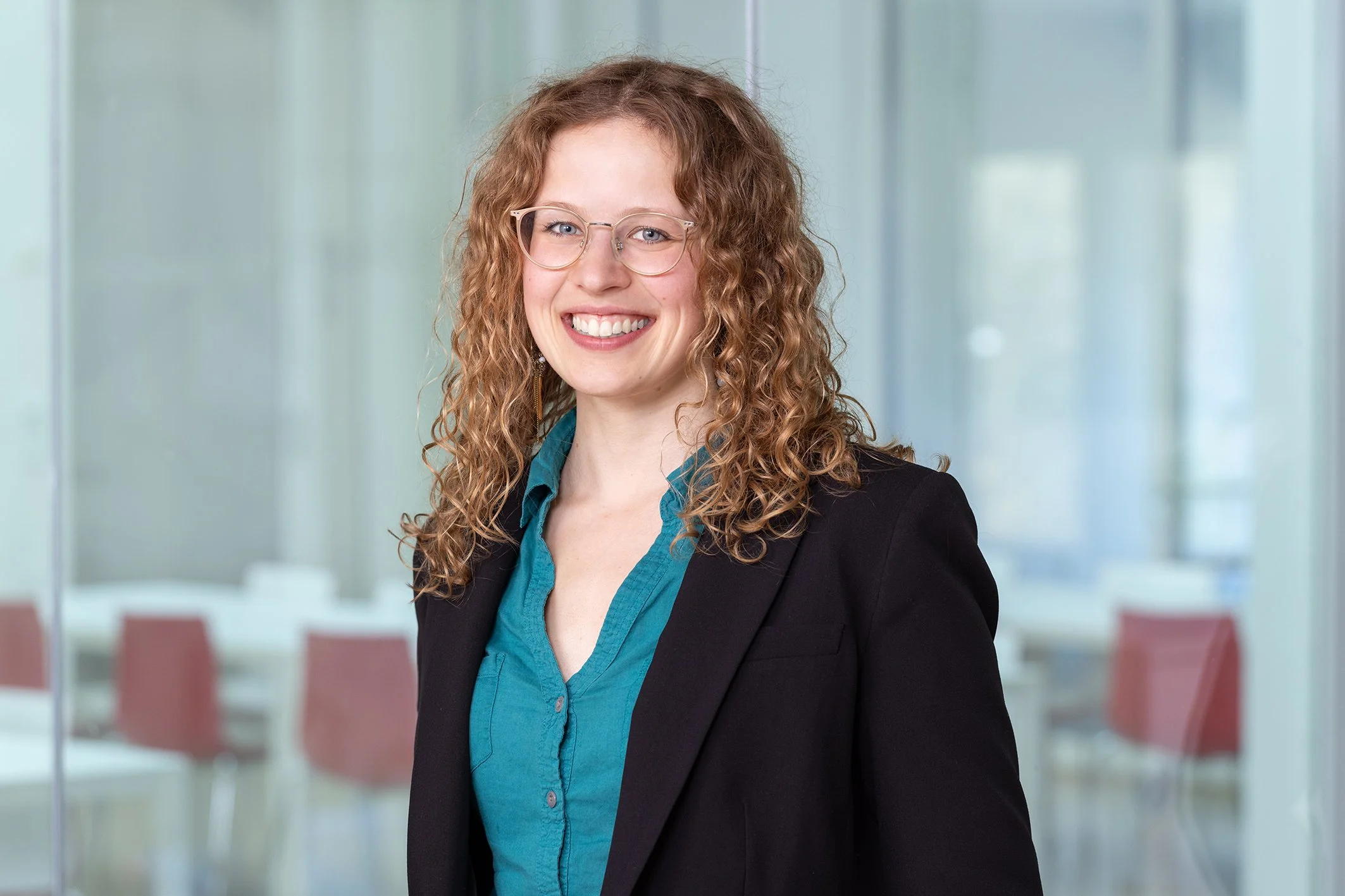 Lächelnde Frau mit Locken, Brille und dunklem Blazer im Büro.