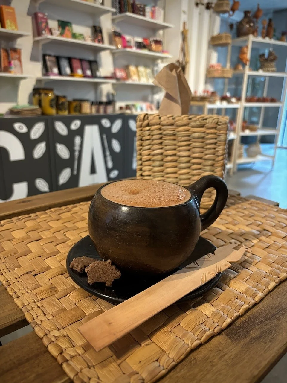 Taza de cerámica con bebida caliente, acompañada de galletas, en un lugar que parece una cafetería, sobre una alfombra de cuerda natural, con fondo de estantes con productos y decoración de madera y plásticos.