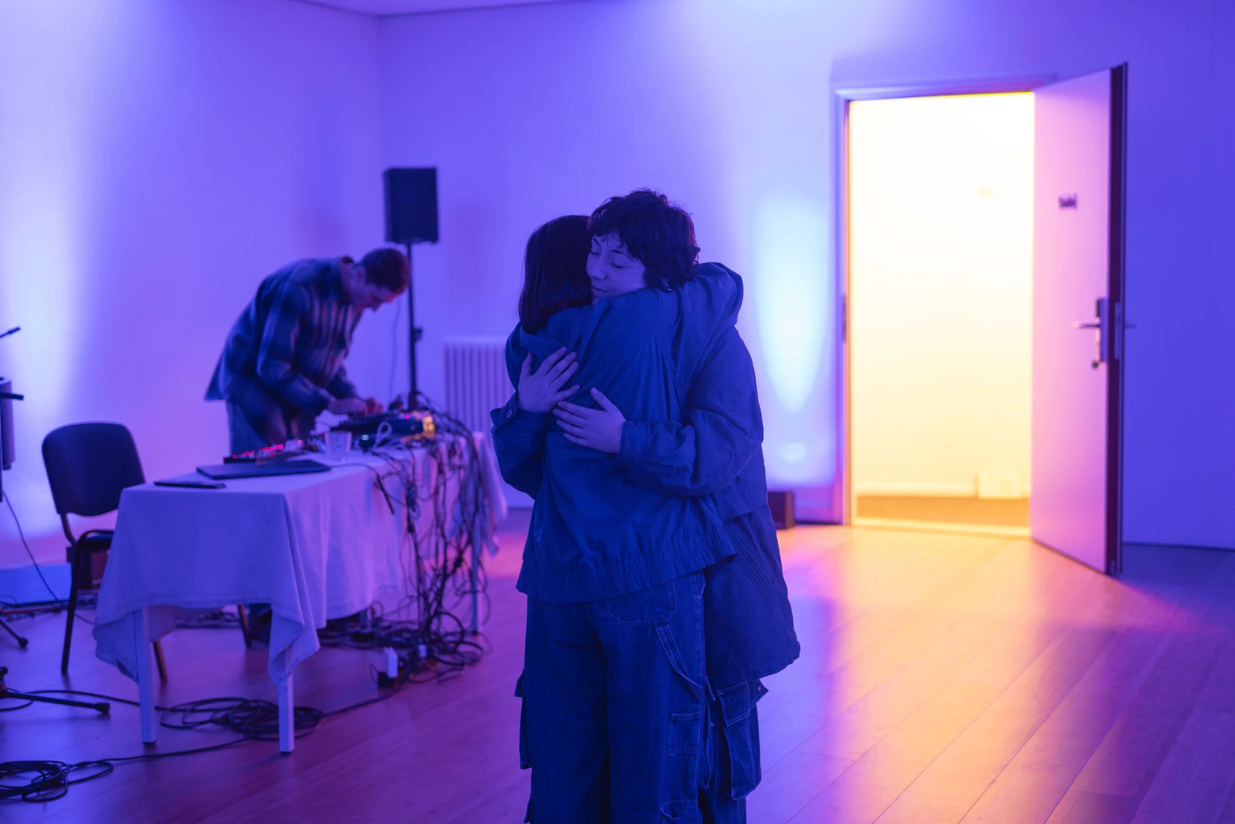 Two people hugging in a room with purple and yellow lighting, with a DJ on a table in the background.