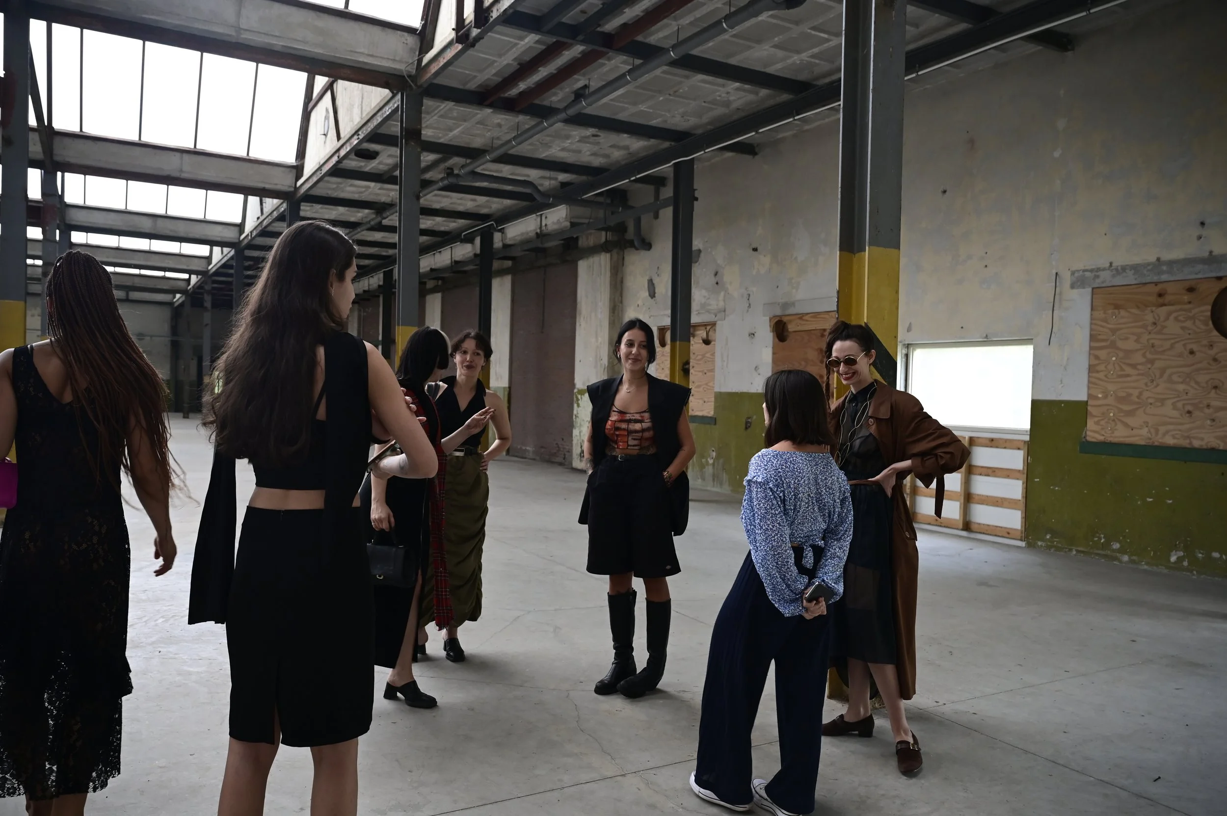 A group of six women in fashionable clothing standing and talking inside an industrial-style space with exposed beams, large windows, and a concrete floor.