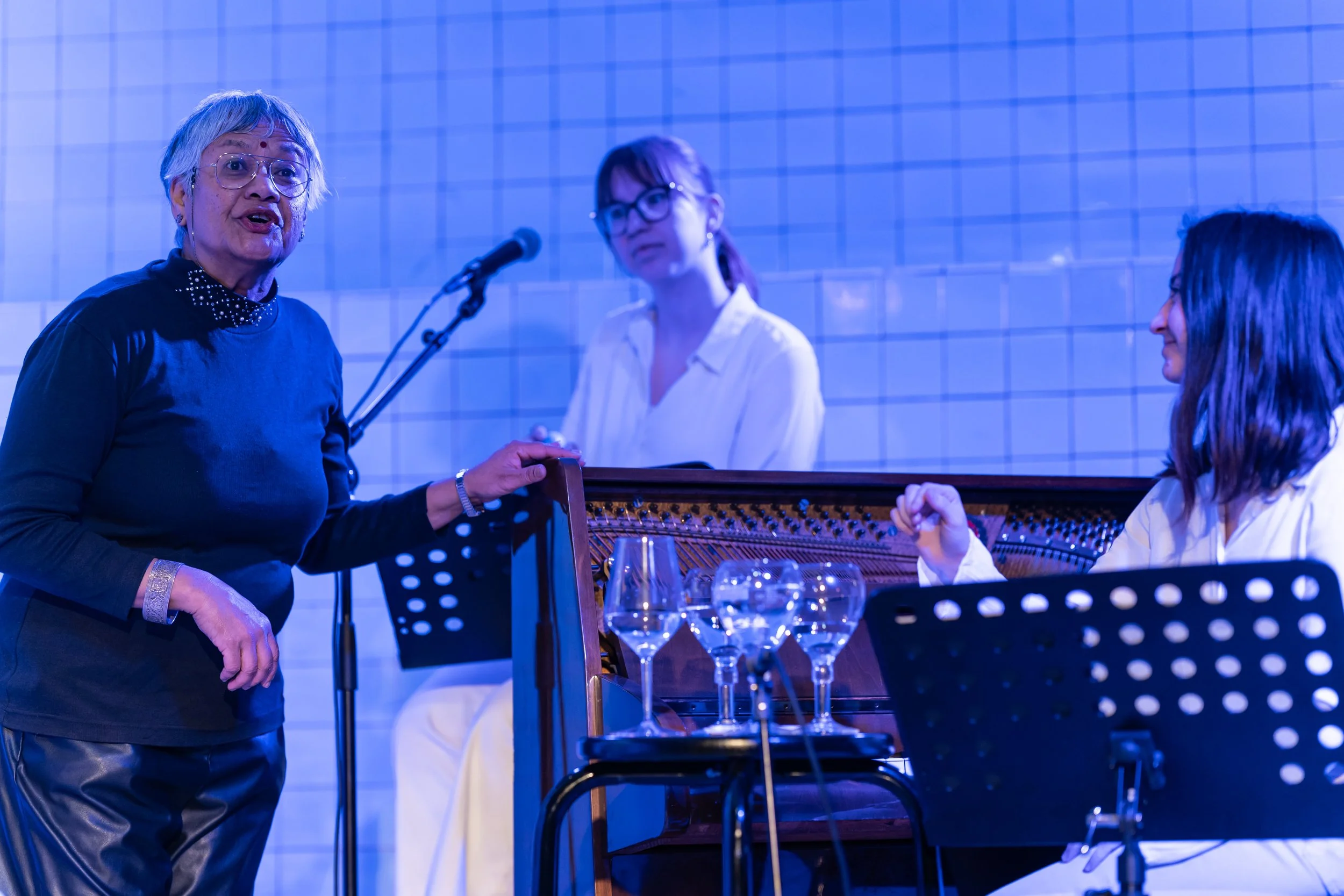 An elderly woman speaking into a microphone with two women listening, one playing the piano, in a room with white tiled walls.