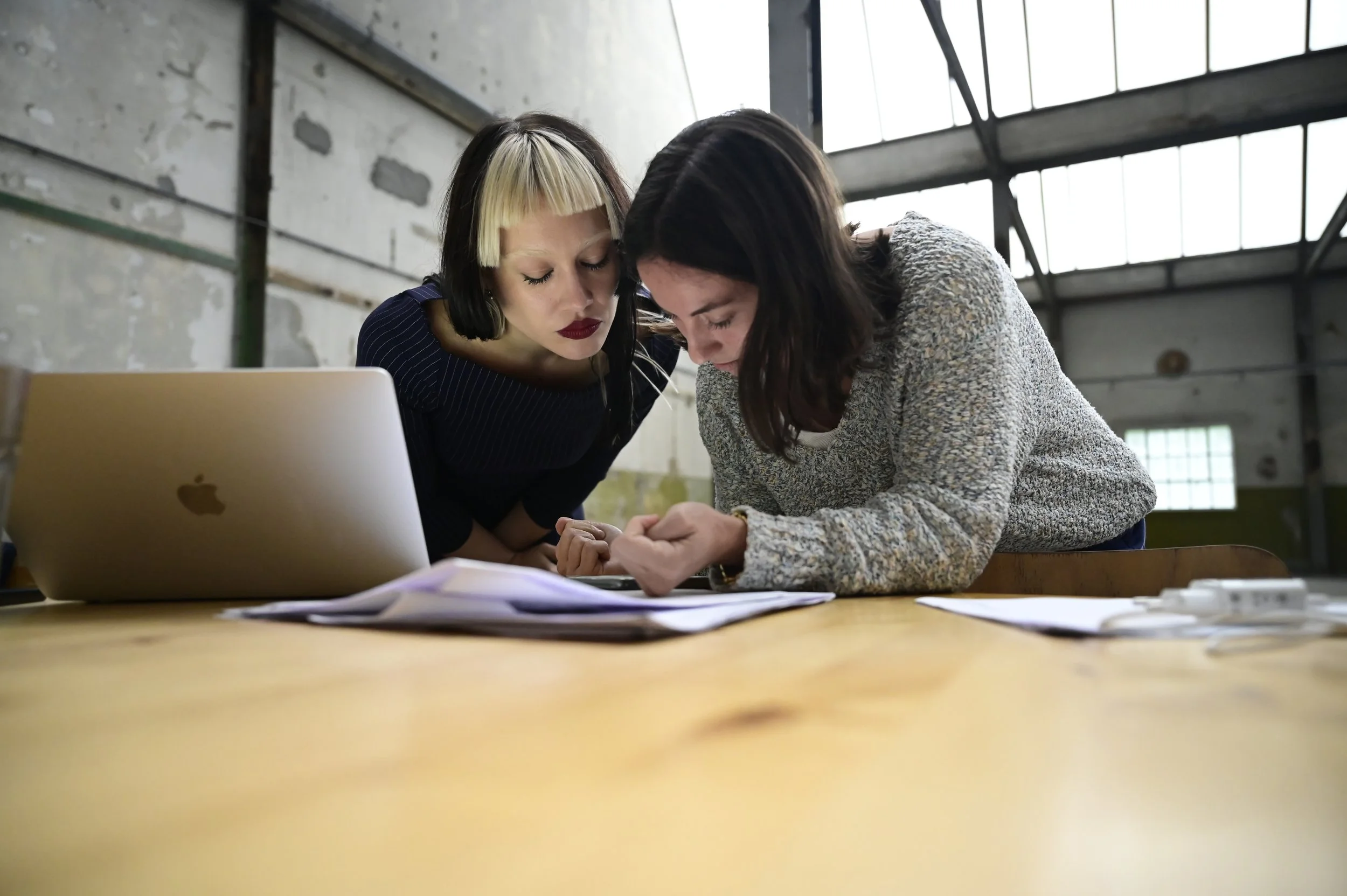 Two women are closely examining papers on a wooden table in an industrial-style room with large windows and exposed walls, with a silver Apple laptop nearby.