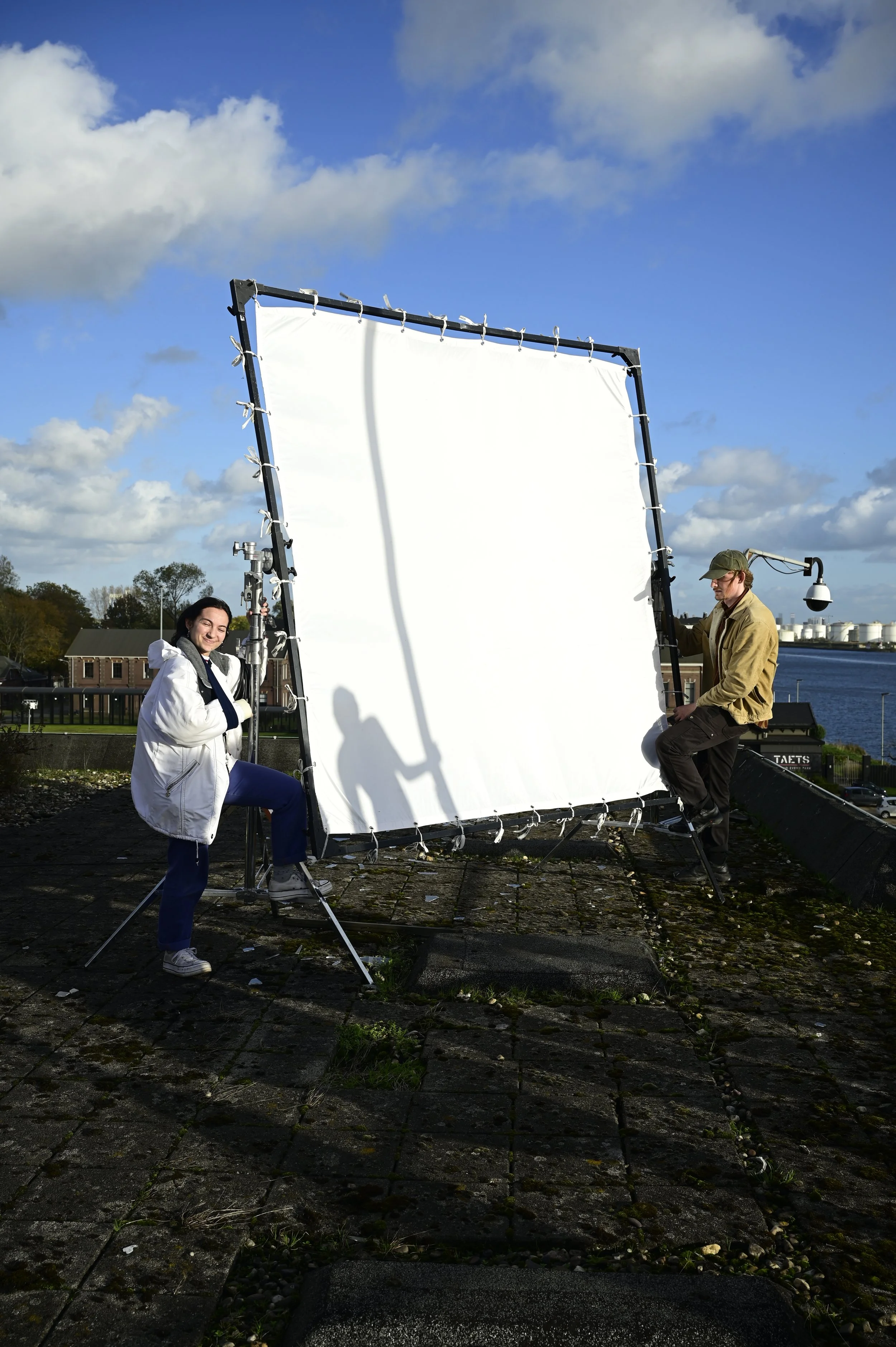 Two people adjusting a large white screen outdoors on a rooftop during daytime, with water and industrial buildings in the background.