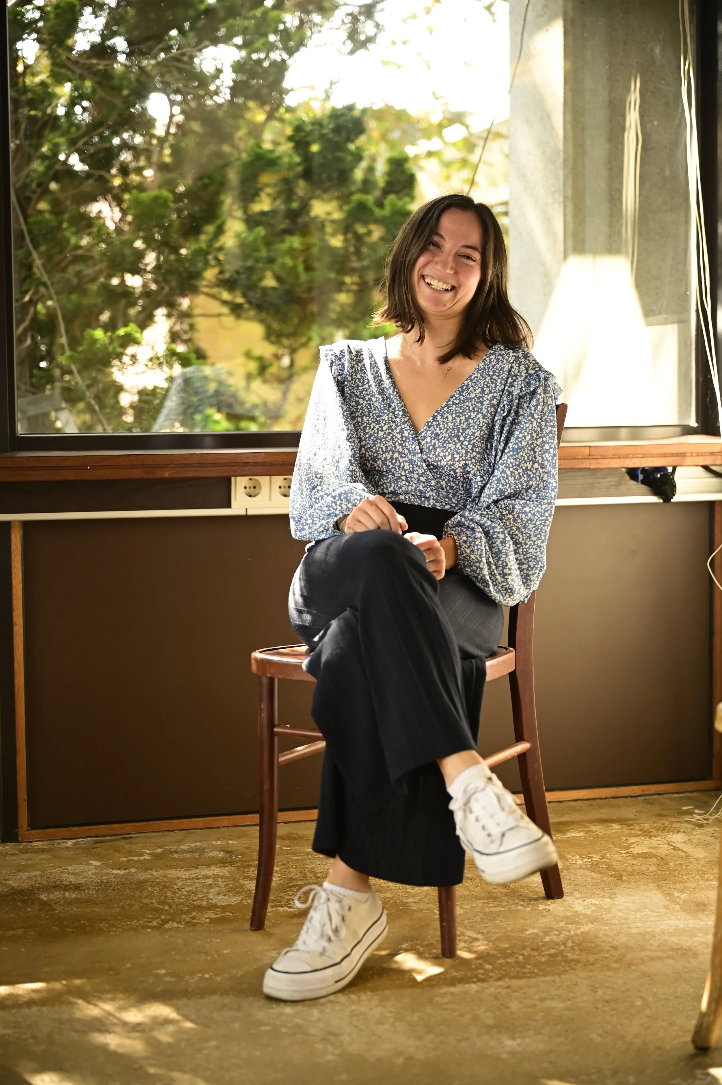 A young woman with brown hair smiling and sitting on a wooden chair inside a room near a window with greenery outside, wearing a blue and white floral blouse, black pants, and white sneakers.