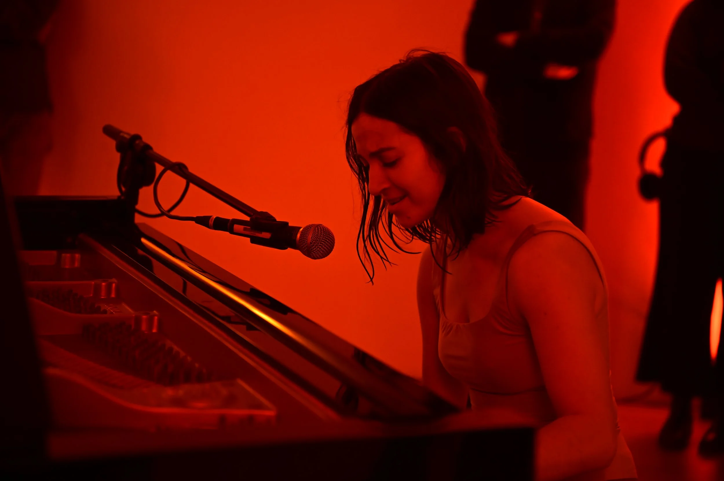 A woman with dark hair singing or recording while playing the piano in a dimly lit, red-orange room, with a microphone positioned near her mouth.