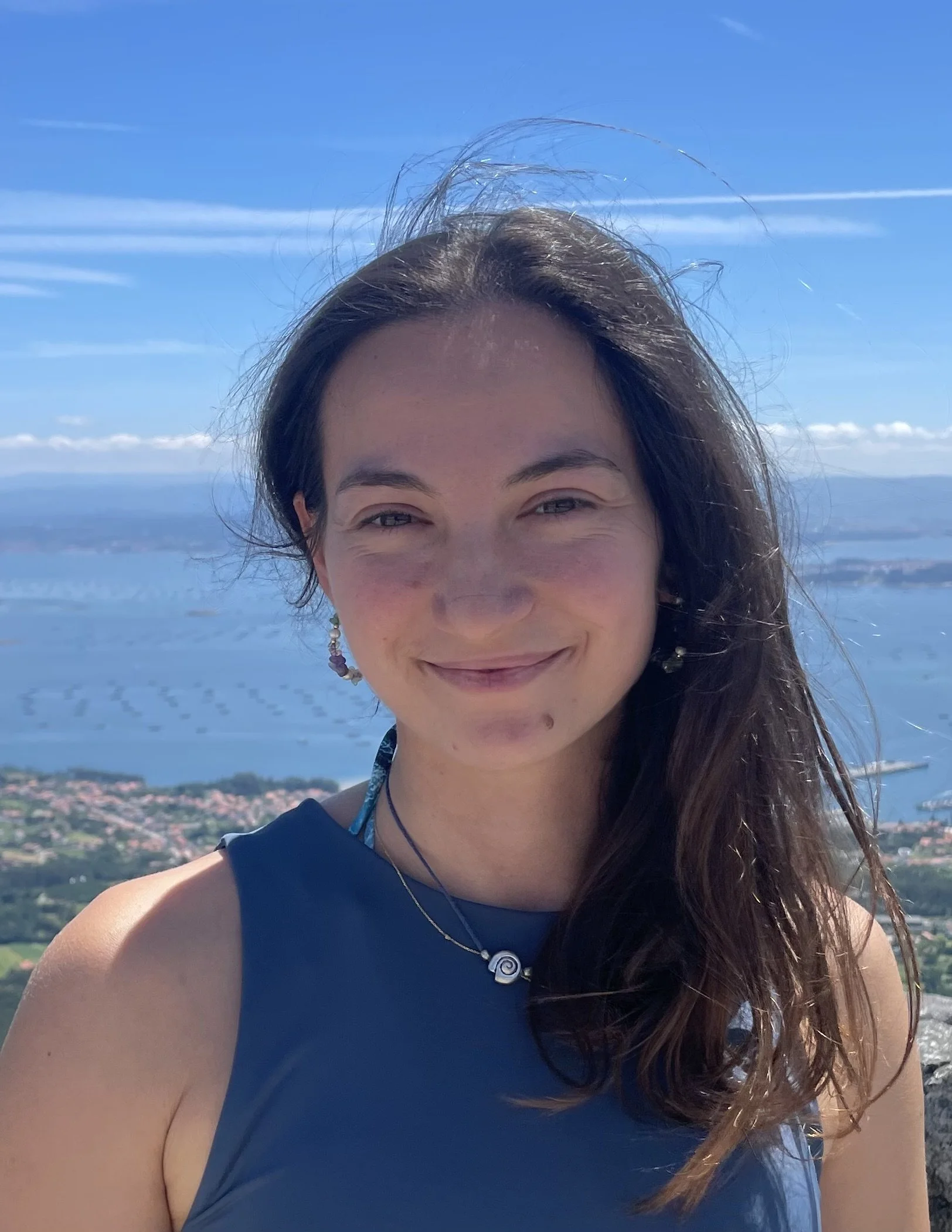 A young woman with long brown hair smiling outdoors against a backdrop of water and land, wearing a sleeveless blue top and jewelry.