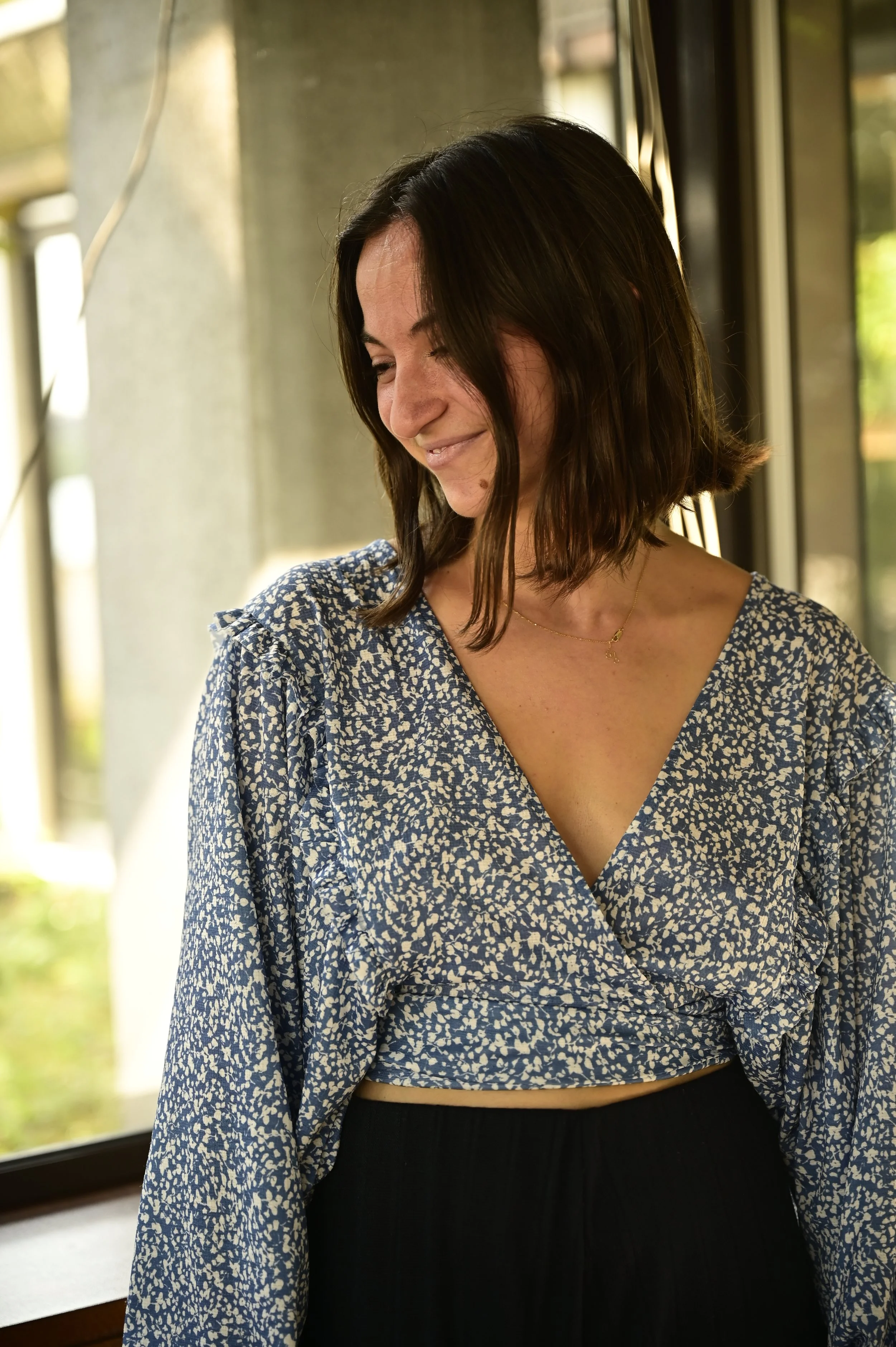 A young woman with shoulder-length dark hair, wearing a blue and white patterned wrap top, smiling and looking downward near a window.