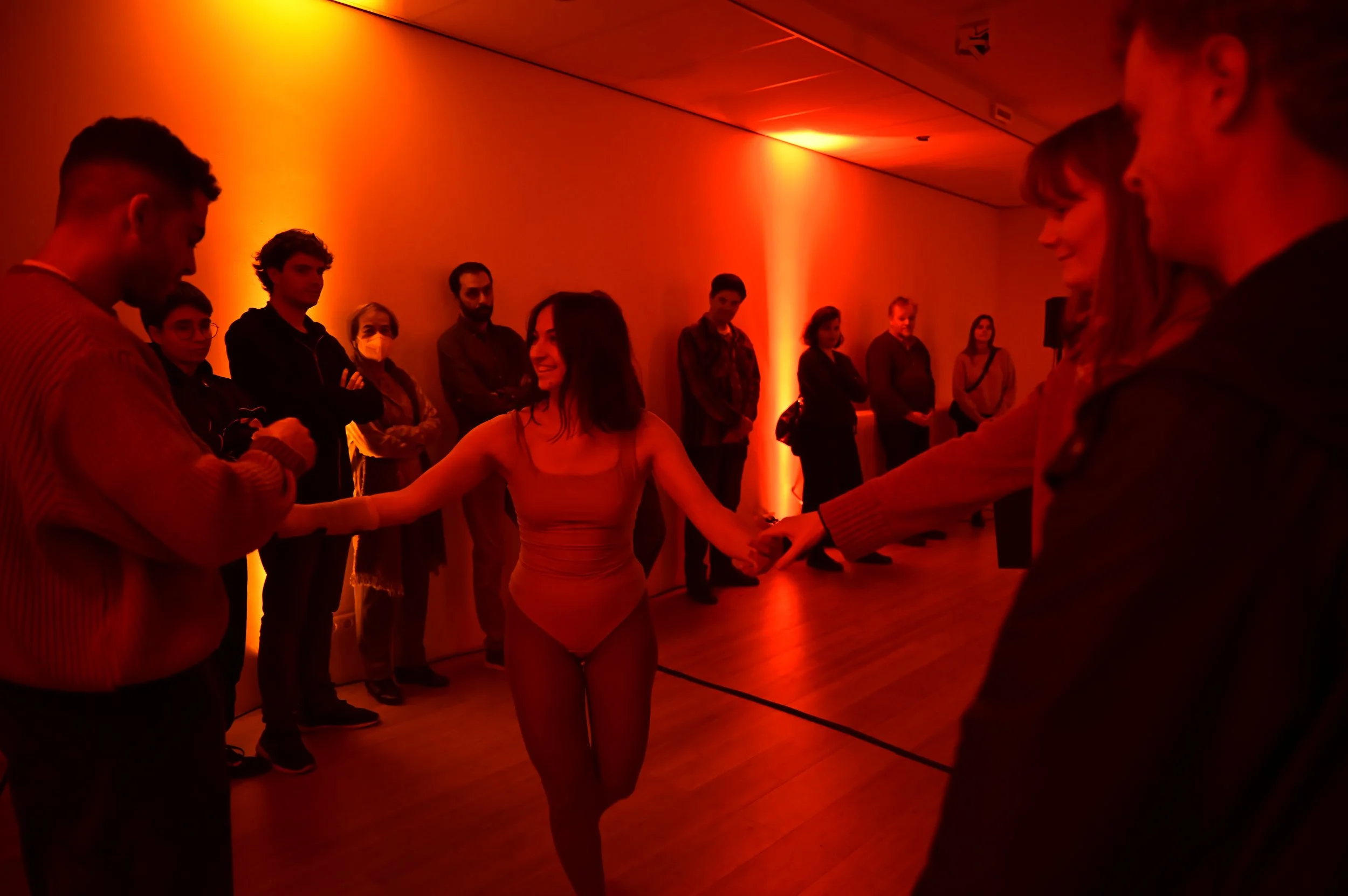 A woman in a tan leotard and fishnet tights dances in a dimly lit room with orange lighting, holding hands with a man. A group of people watches her from the side, standing against the wall.