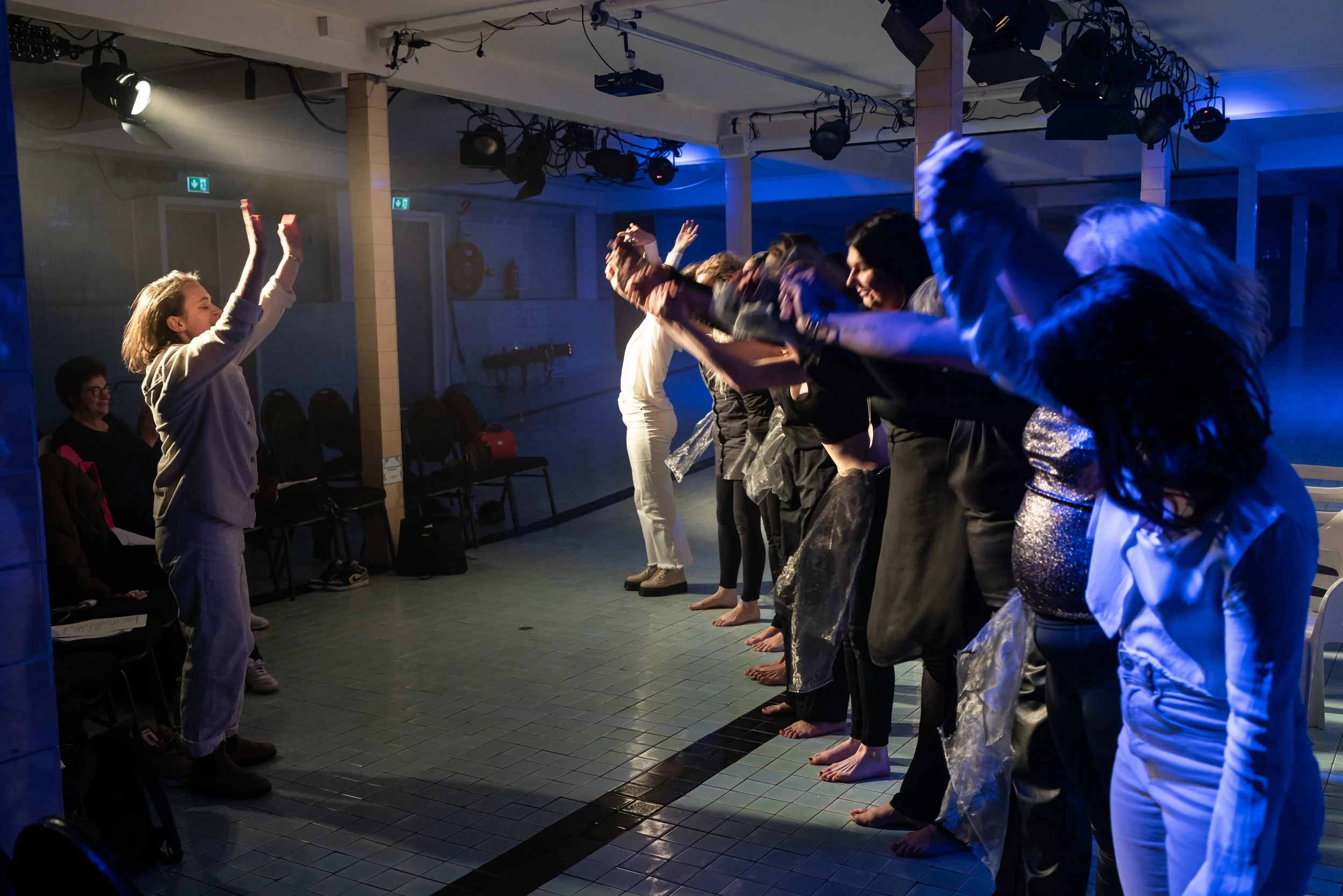 A group of people standing in a line, raising their arms in the air, with one person at the front appearing to lead or conduct them, in a room with stage lighting and blue hues.