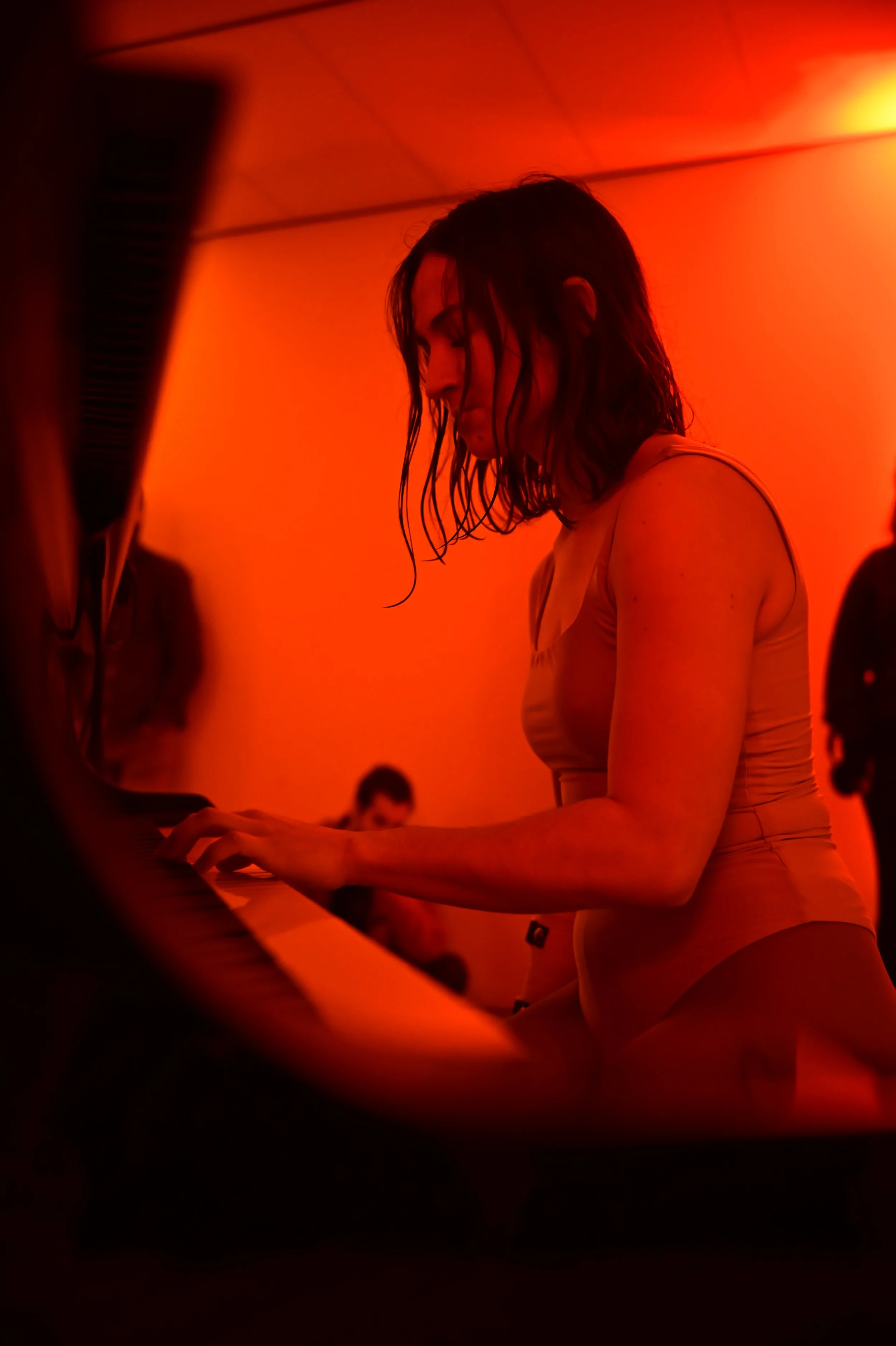 A woman with wet hair playing the piano in a dimly lit room with red lighting, piano and several blurred figures in the background.