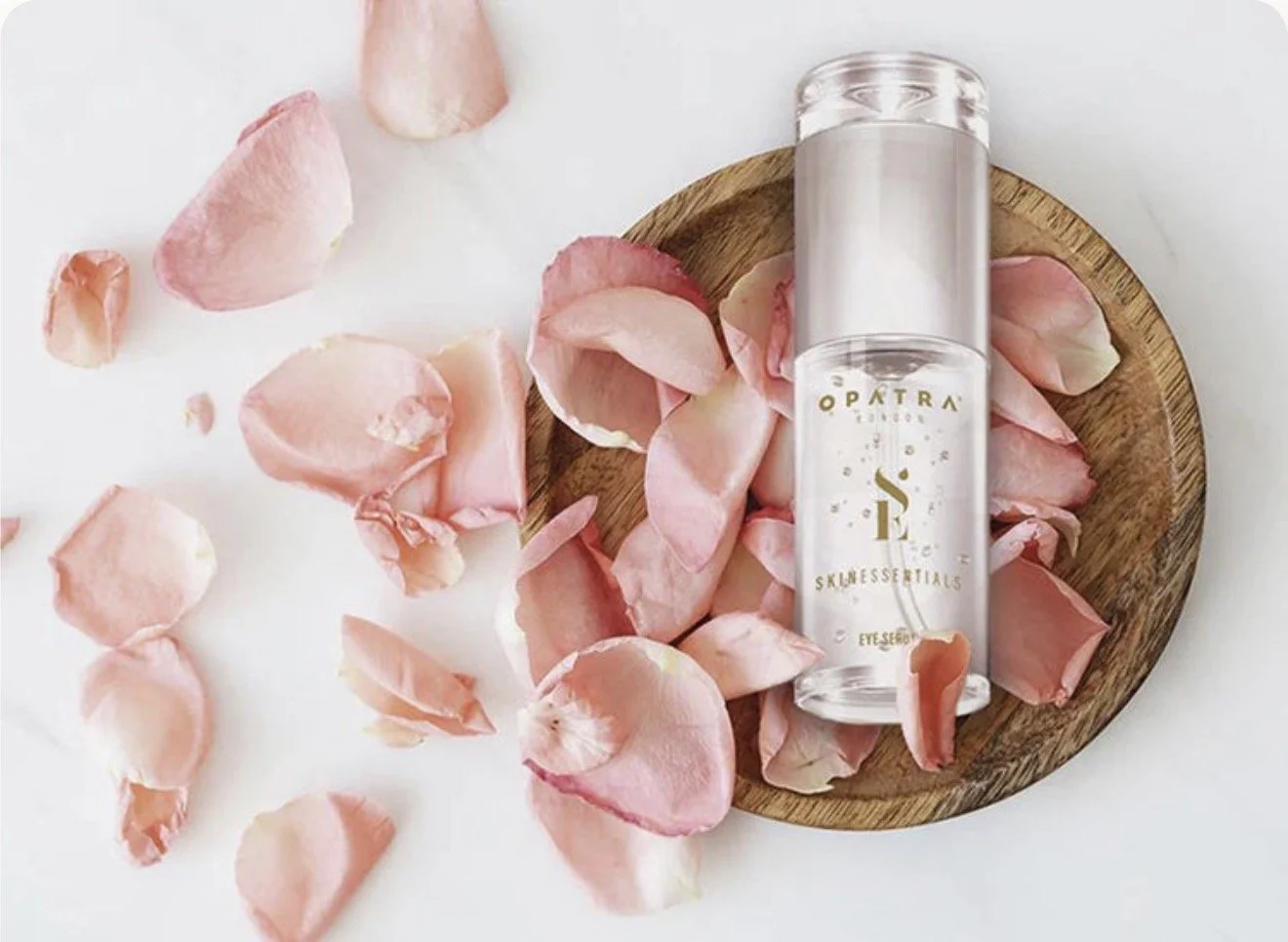 A clear cylindrical skin serum bottle on a small round wooden board surrounded by pink rose petals on a white surface.