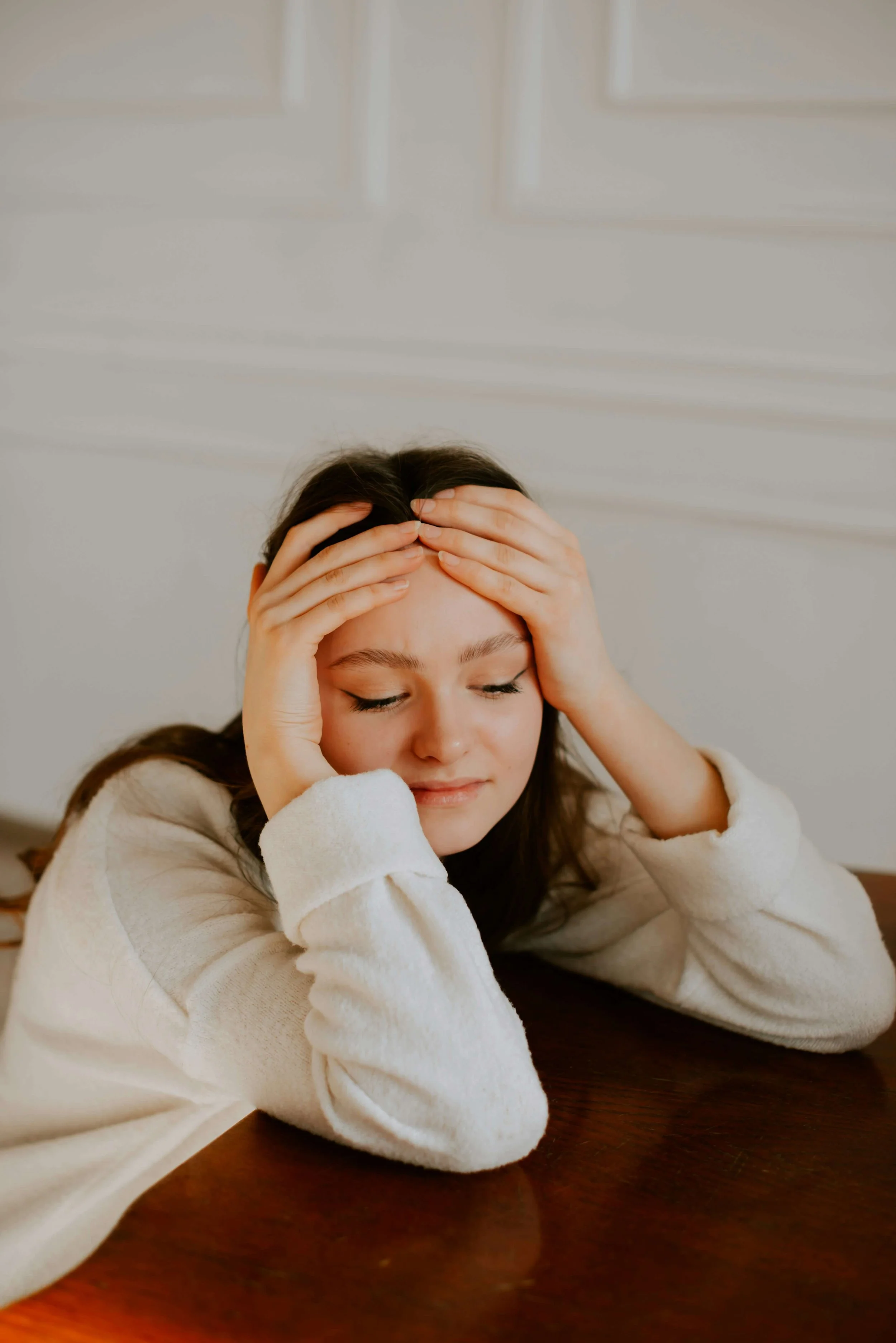 A woman in a white sweater looks down, holding her forehead