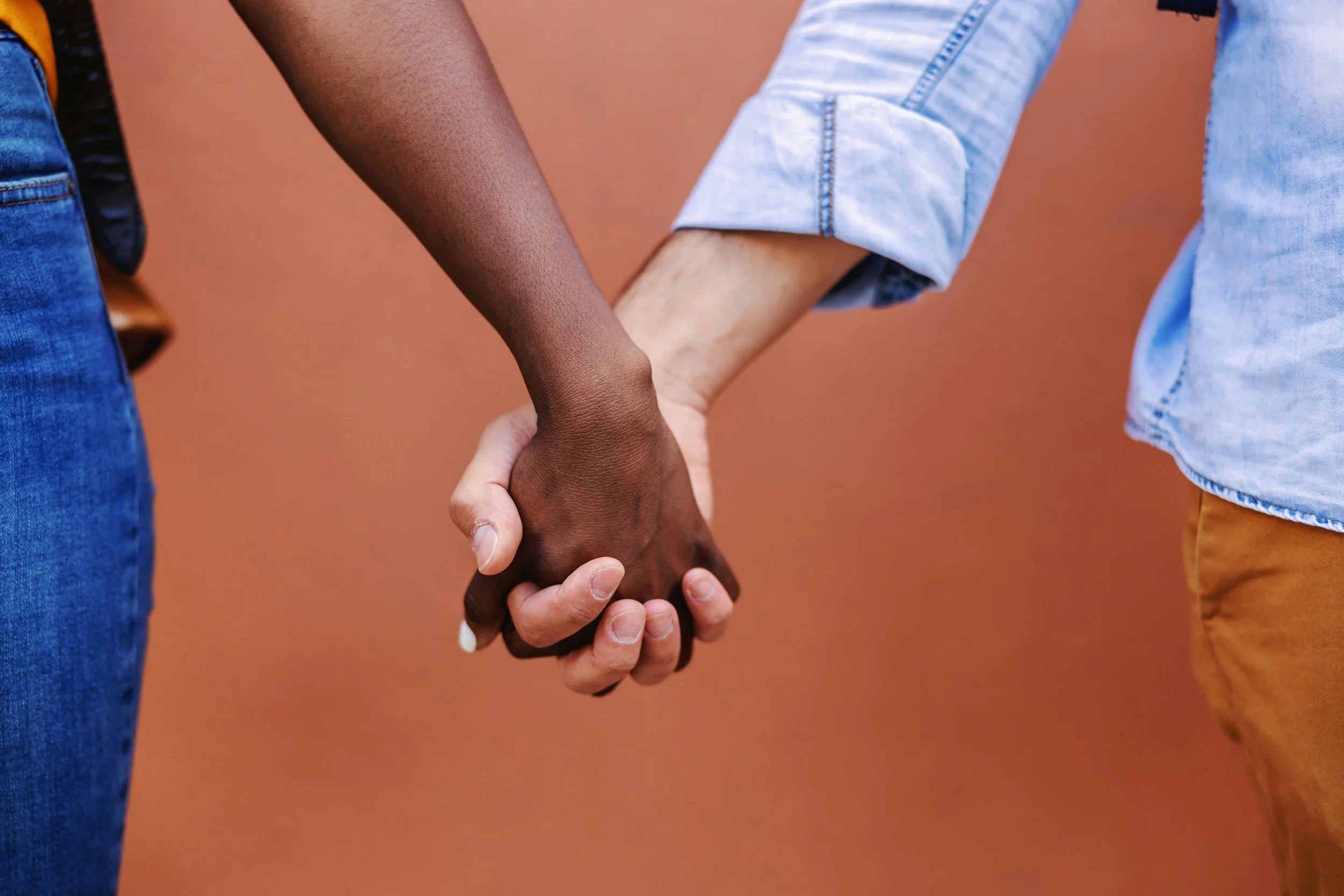 A Black person and a white person hold hands before a muted orange background