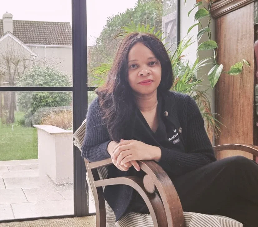 A woman with shoulder-length black hair, wearing a black sweater, sitting in a wooden chair indoors next to a large window, with a garden visible outside and a potted plant behind her.