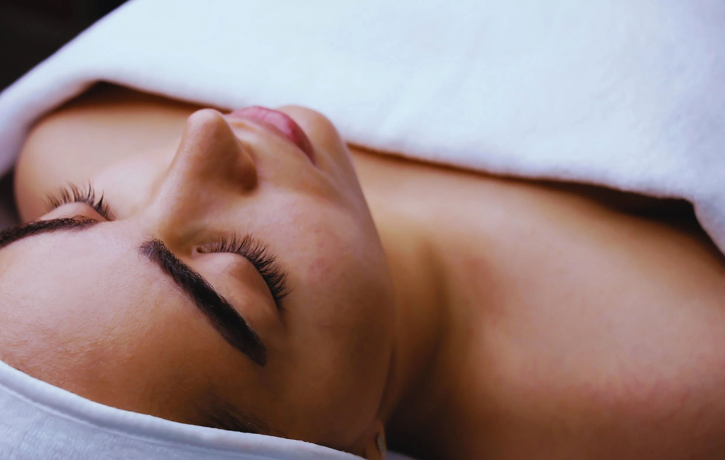A woman lying down with eyes closed, covered with a white towel or sheet.