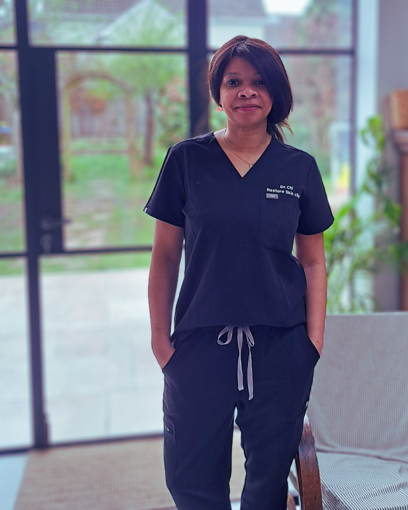 A woman wearing black medical scrubs stands indoors near large windows with greenery outside. She has shoulder-length dark hair and is smiling gently, with hands in her pockets.