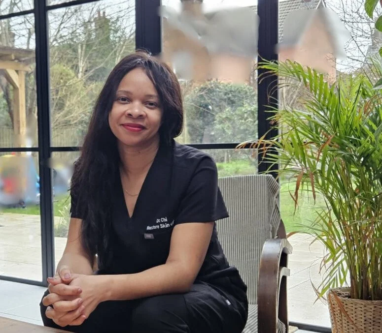 A woman with long dark hair, wearing a black uniform, sitting at a table next to a large potted plant, inside a room with glass windows and a view of the outdoors.