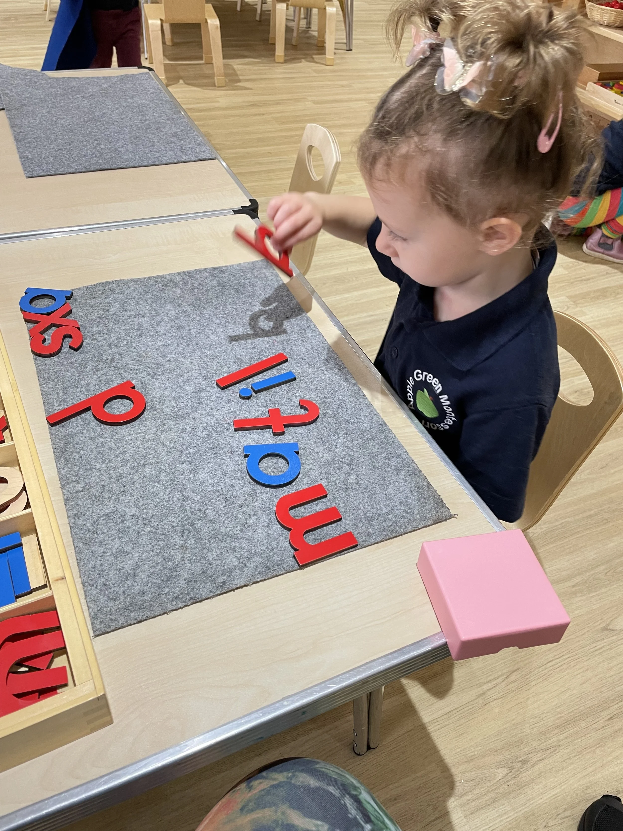 A young girl with curly hair tied with pink hair ties, wearing a dark blue shirt, is sitting at a table arranging colorful foam letters spelling the word 'mats' on grey felt. There are additional foam letters and a pink box on the table.