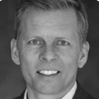 Close-up of a man with short light hair, smiling, in black and white.