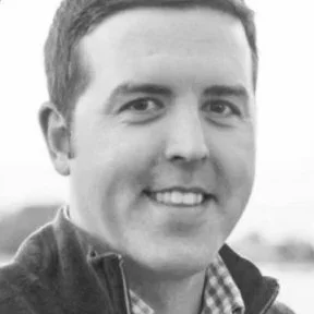 Black and white portrait of a smiling man with short hair, wearing a jacket and a checkered shirt underneath.