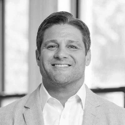 Black and white portrait of a smiling man in a suit, with a blurred office background.