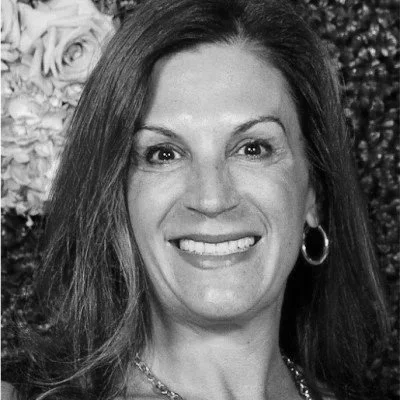 A woman smiling with long hair, wearing hoop earrings and a necklace, with a floral background in black and white.