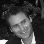 A smiling man with dark hair and a light shirt in an outdoor setting with trees in the background.