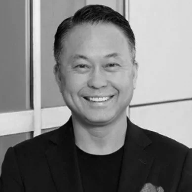 Black and white photo of a smiling man in a blazer and t-shirt, standing against a geometric background.