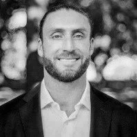 Black and white photo of a man with a beard and short hair, wearing a suit jacket and a collared shirt, smiling outdoors.