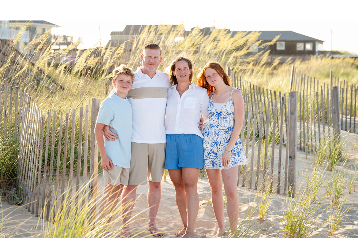 Family of four standing on a sandy beach path with dunes and grass, smiling at the camera, sunny weather