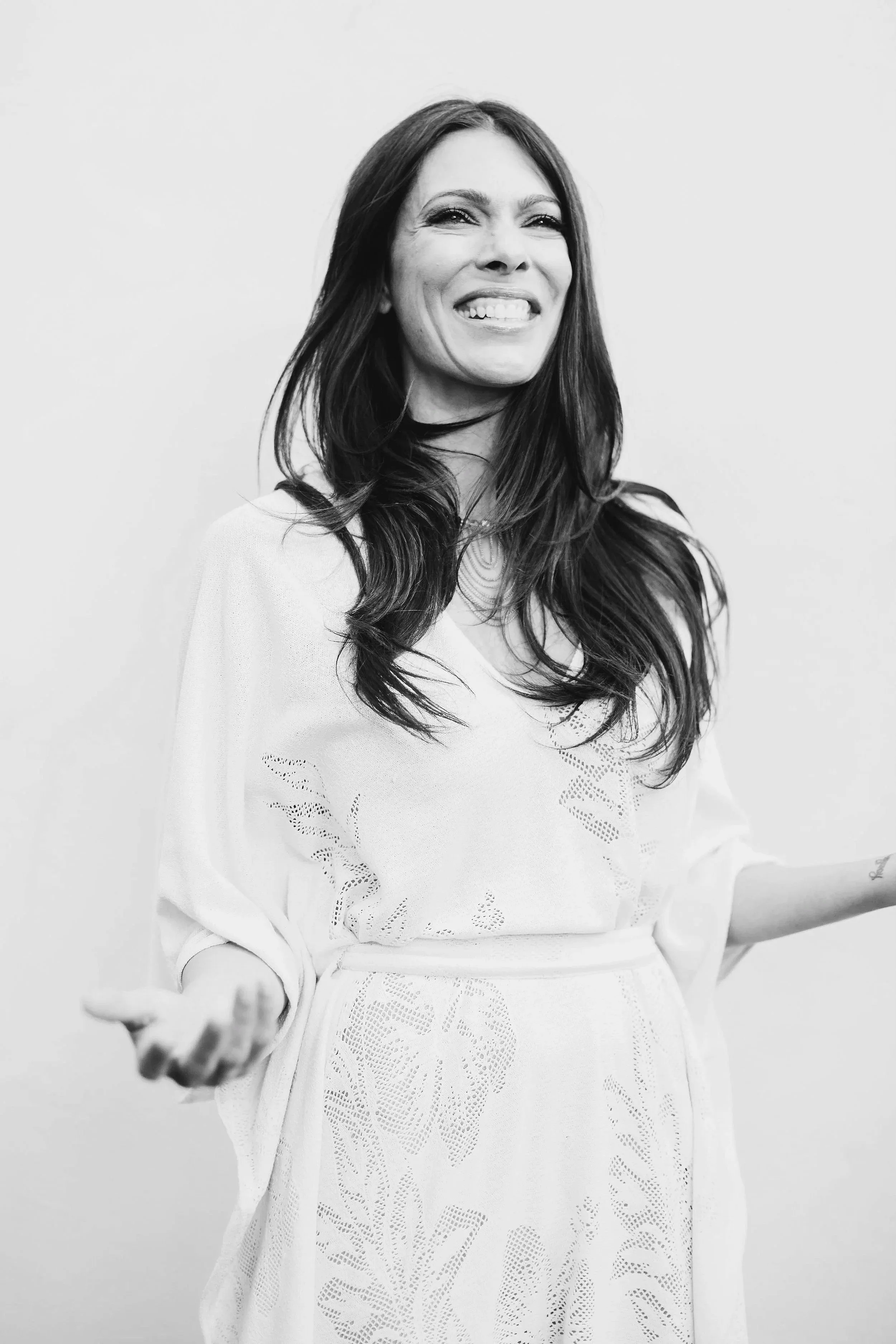 A smiling woman with long dark hair, wearing a light-colored dress with floral cutout details, standing against a plain background. | Michele Lovely | The Birthday Portal | Tedx  | Global Idea Search 2025