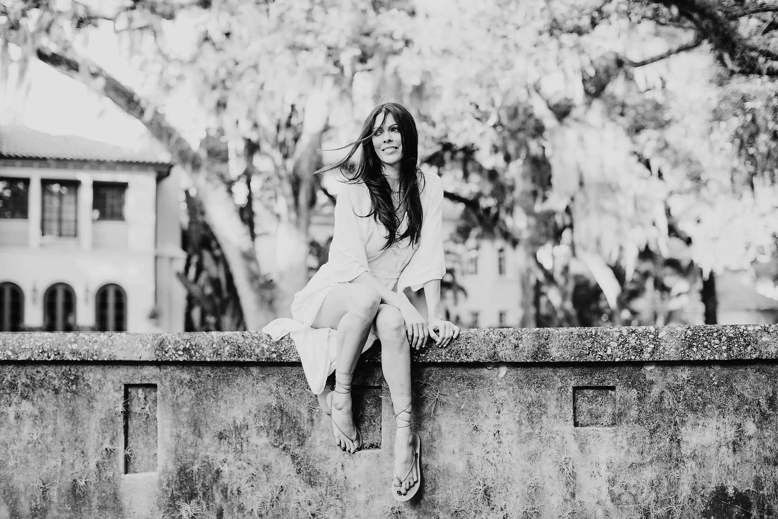 A woman in a light-colored dress sitting on a stone wall outdoors with trees and buildings in the background, in black and white. | Michele Lovely | The Birthday Portal | Tedx  | Global Idea Search 2025