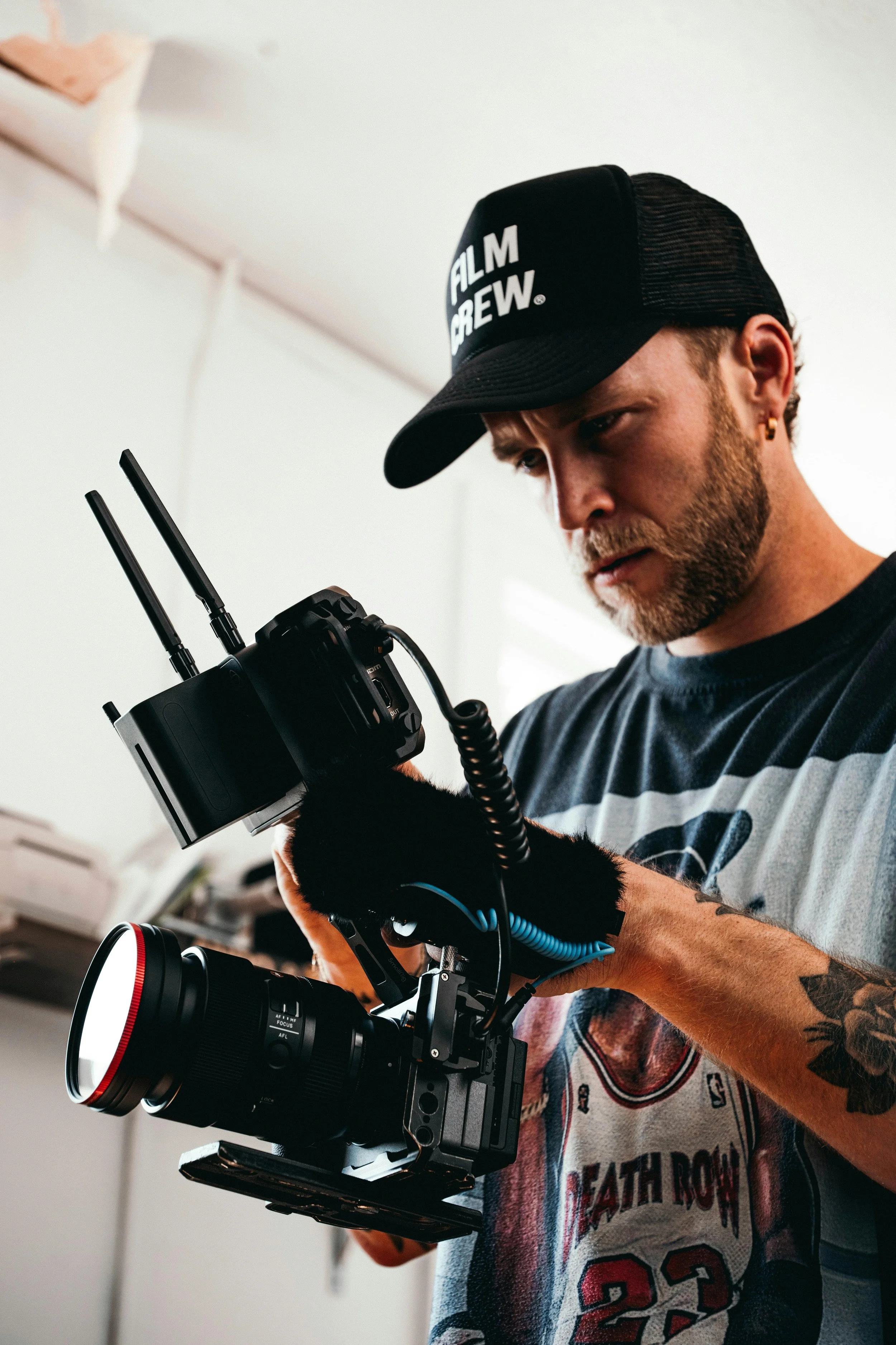 A man with a beard, wearing a black hat with 'FILM CREW' written on it, is adjusting a camera with a large lens. He is indoors, wearing a graphic T-shirt with a sports theme.